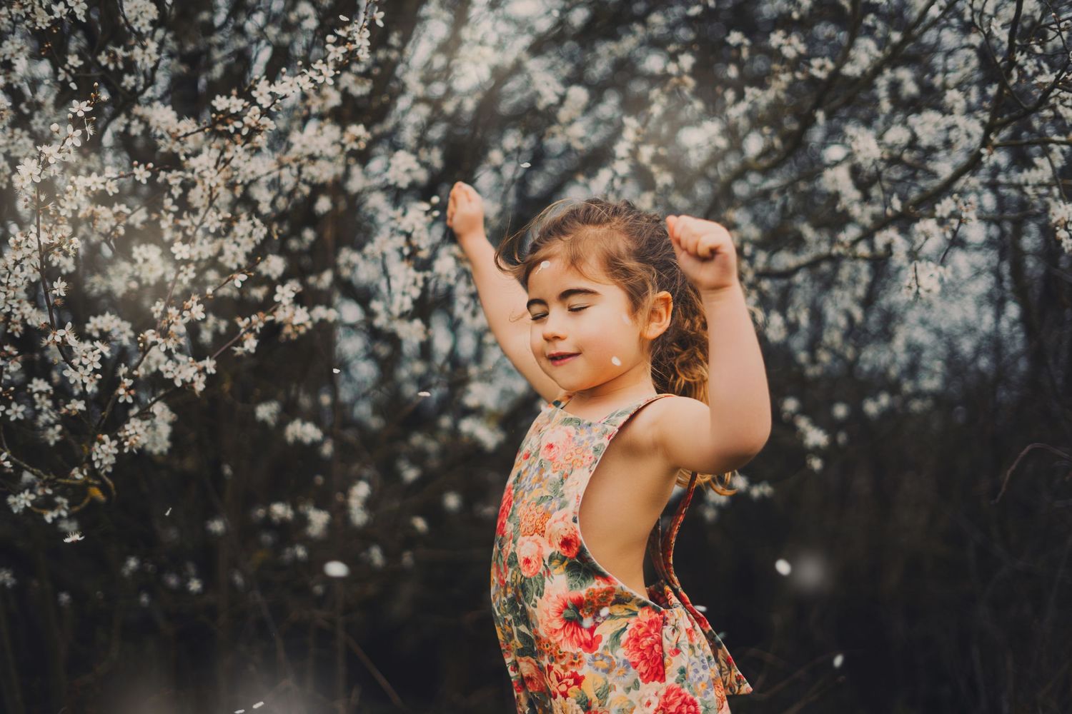Young girl in floral dress dancing with arms raised near white blossoming trees in soft light.