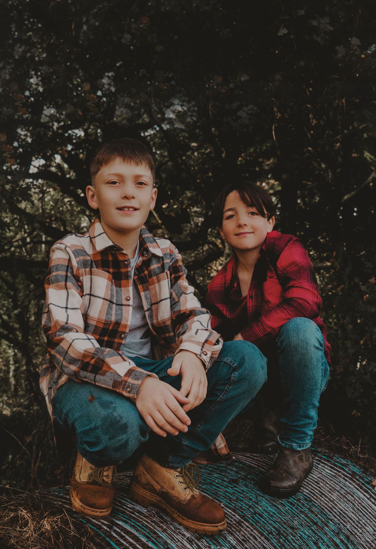 Two boys in flannel shirts and jeans crouching outdoors in a forest setting, smiling at the camera.