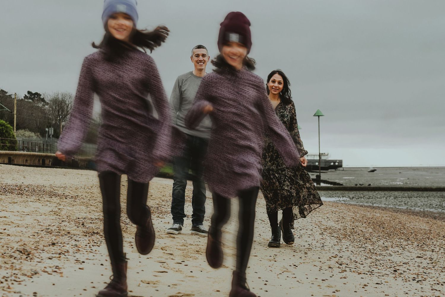 Family of four on a pebble beach, two girls running blurred in foreground, parents smiling behind them.