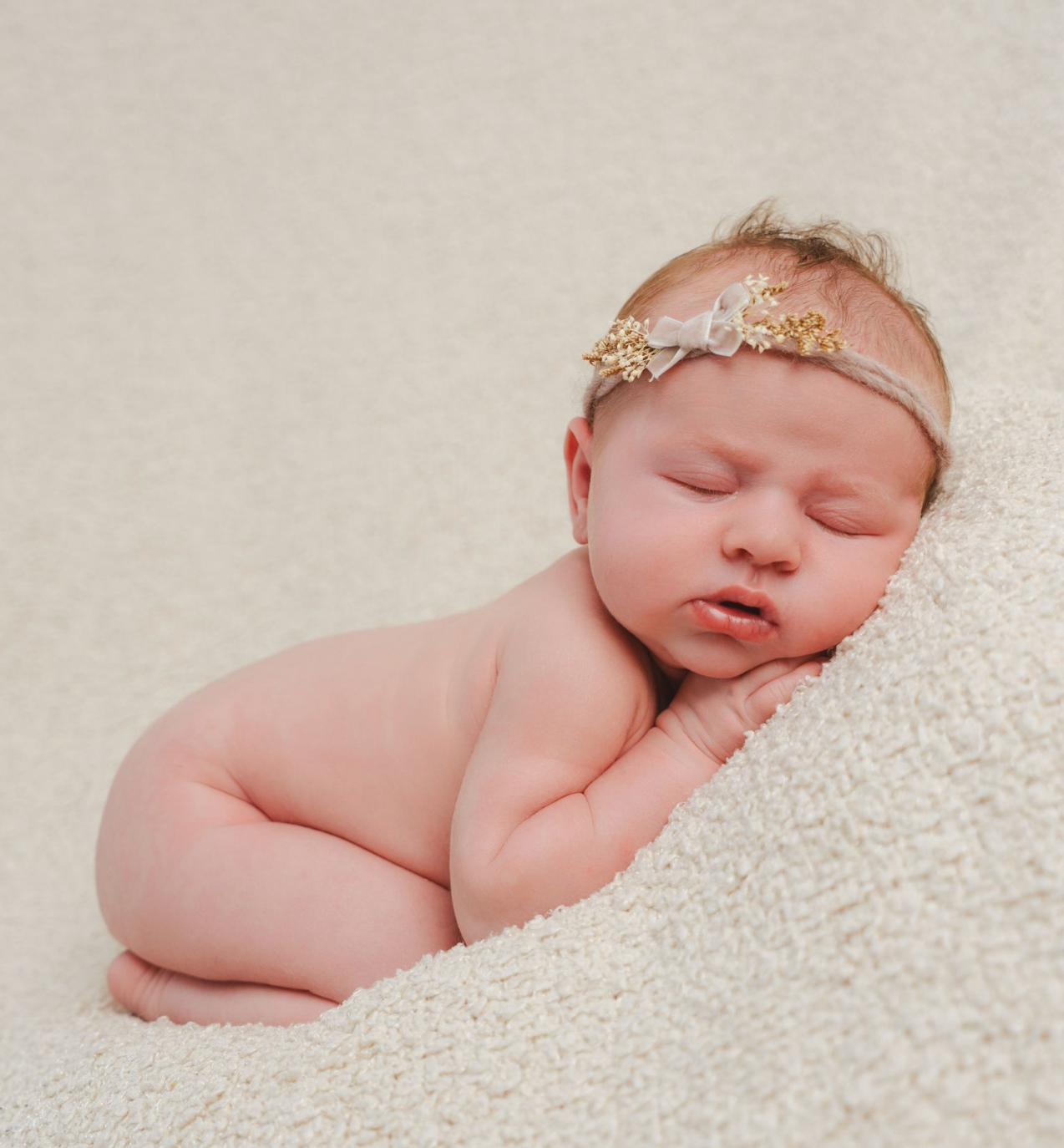 Sleeping newborn baby girl wearing a delicate gold floral headband, posed on a soft cream blanket.