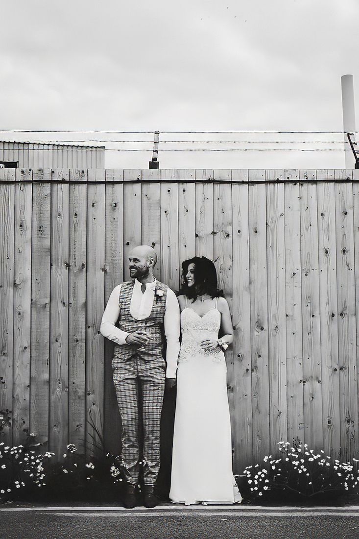 Bride and groom standing against wooden fence in black and white portrait.
