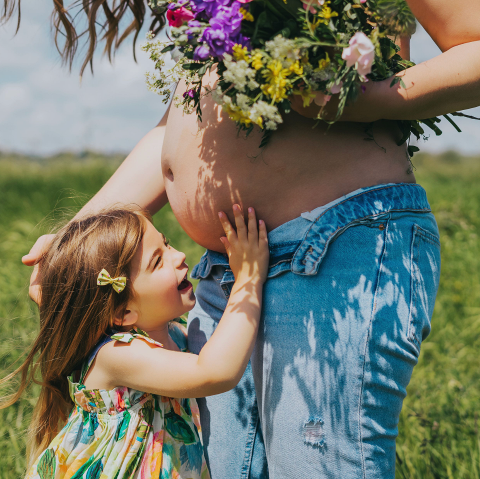 A young girl in a floral dress kisses a pregnant woman's bare belly outdoors in a sunny green field.