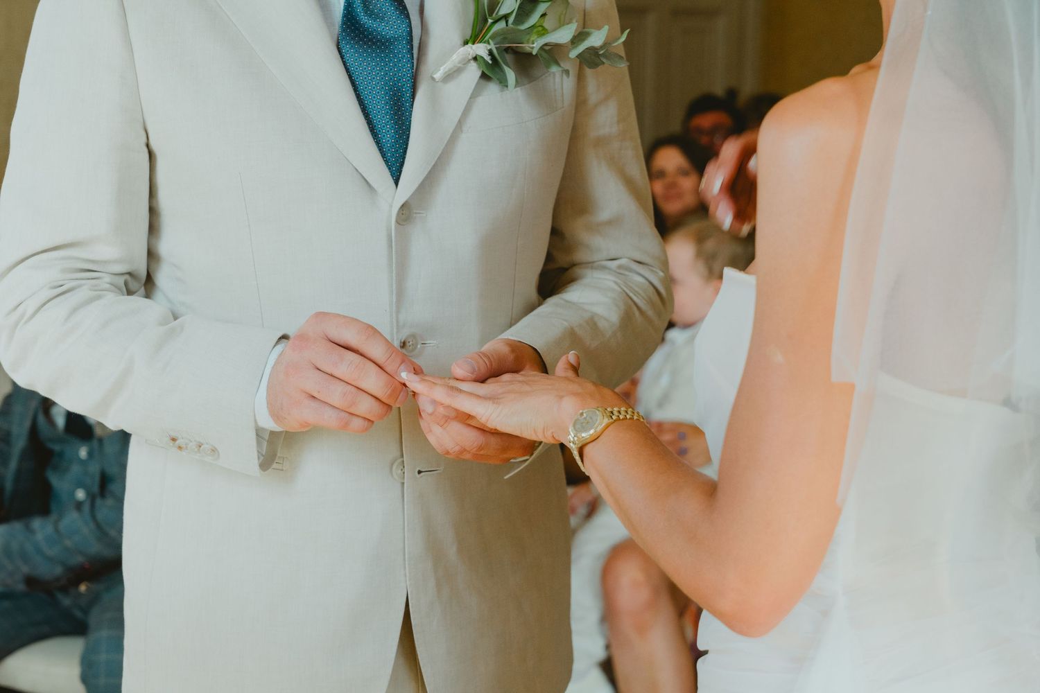 Groom in beige suit placing wedding ring on brides hand during outdoor ceremony exchange of vows.