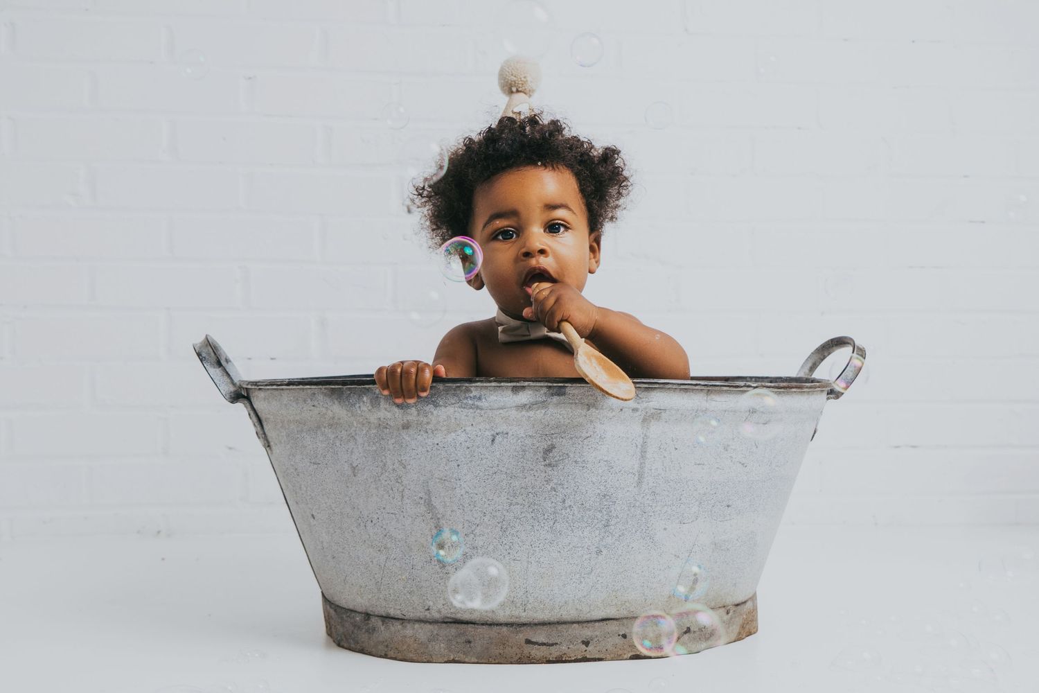 Adorable Black baby sitting in vintage metal tub holding a colorful toy indoors.