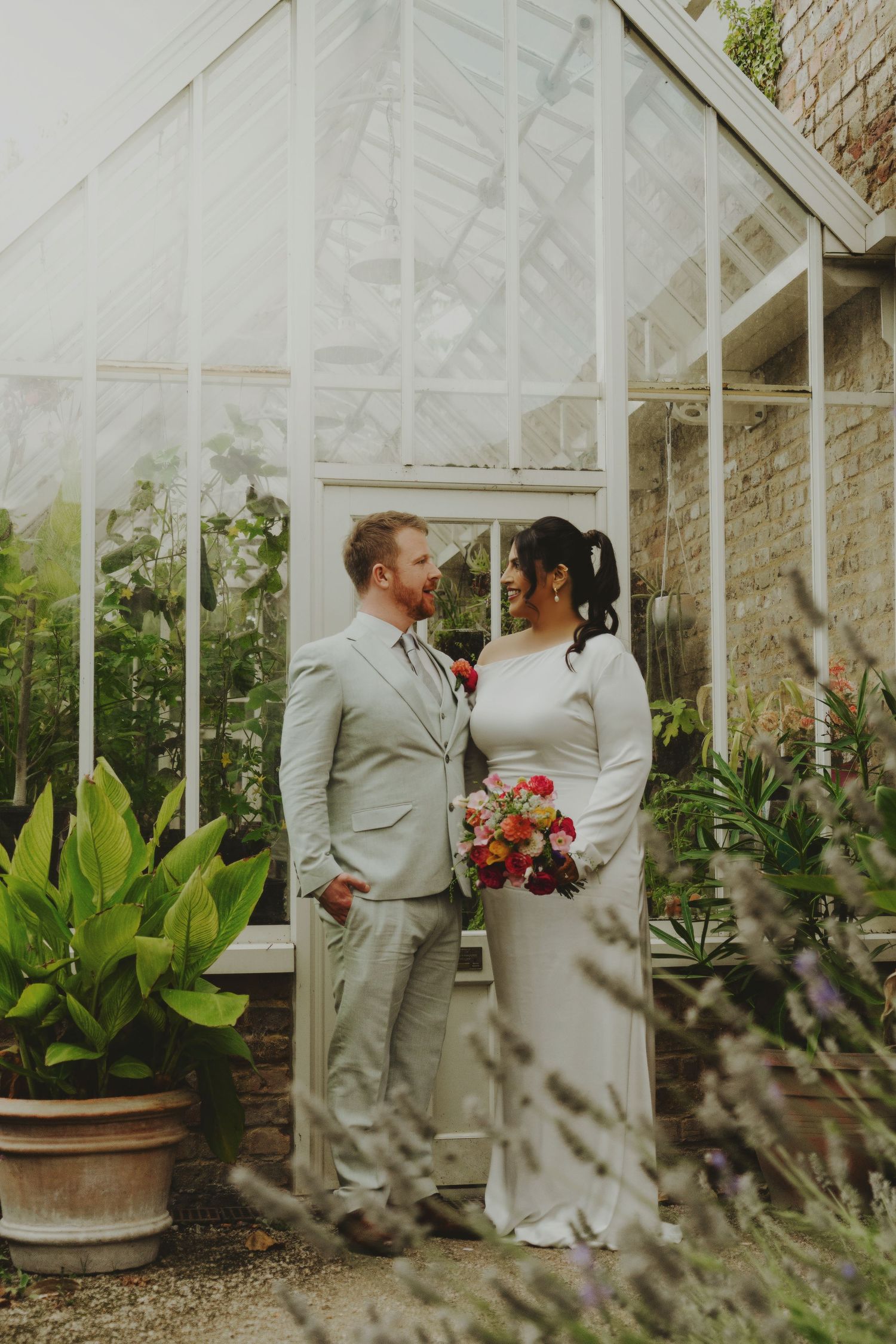 Bride and groom smiling at each other in front of a charming greenhouse with vibrant flowers.