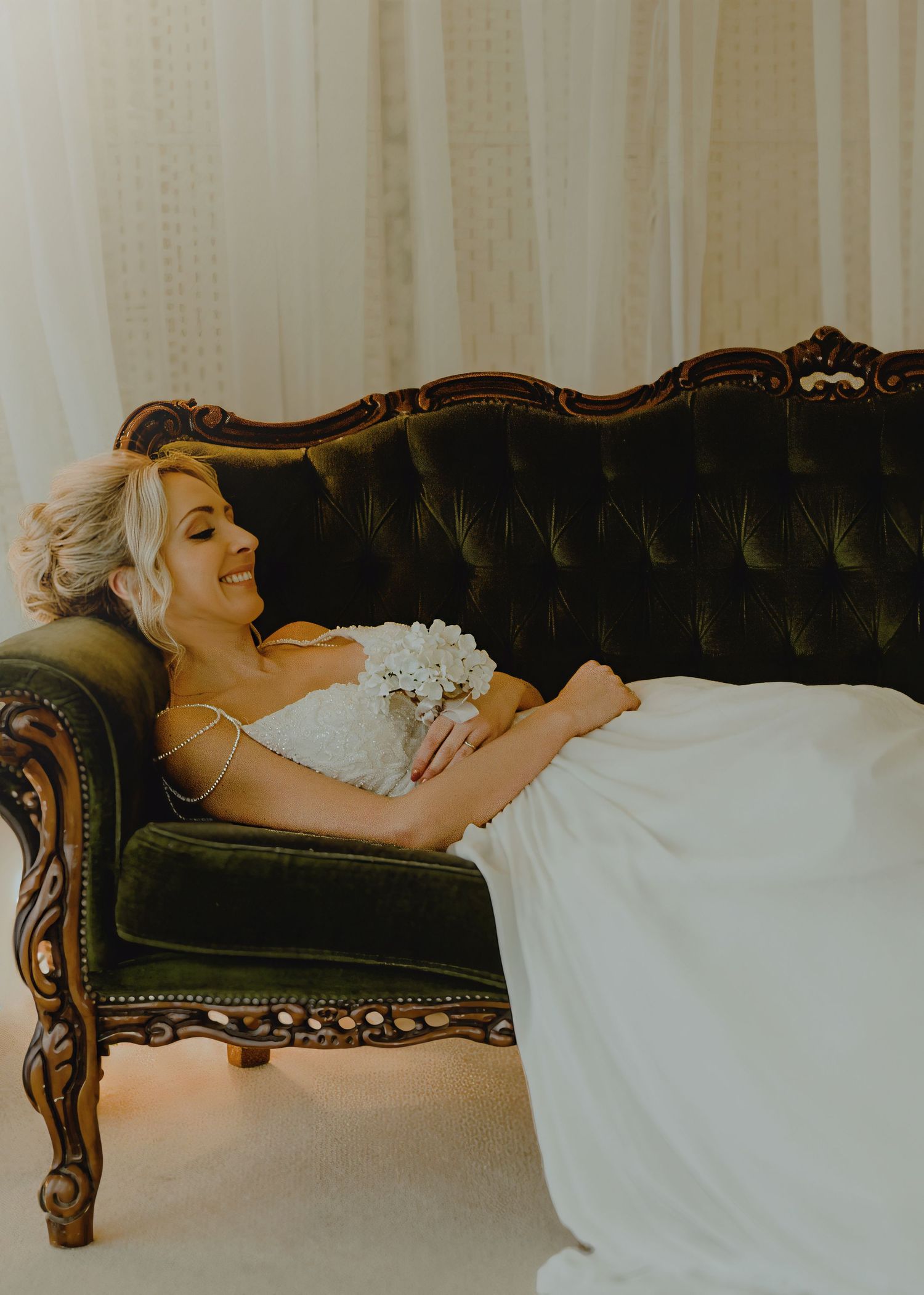 Bride in white gown reclining on ornate green velvet tufted sofa holding bouquet, smiling softly.