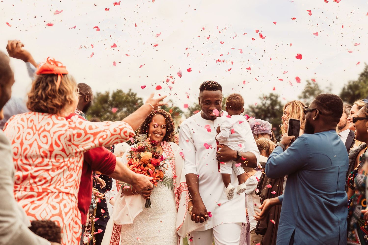 Joyful newlyweds walk through colorful confetti shower surrounded by celebrating wedding guests outdoors.
