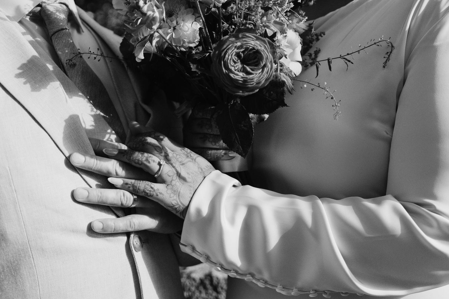 Black and white close-up of wedding rings on clasped hands holding a floral bouquet.