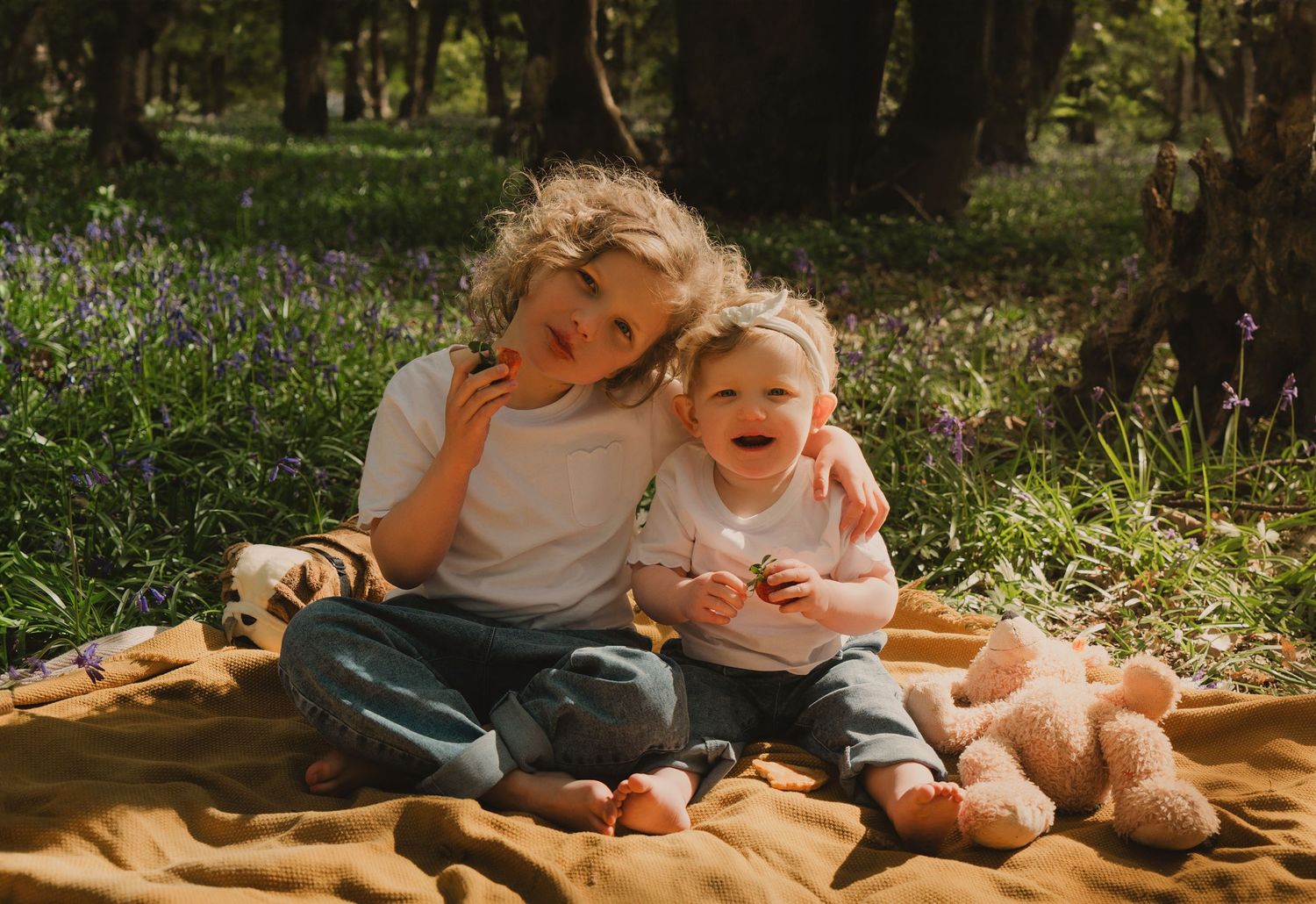 Two young siblings sit together on a blanket in a sunny bluebell forest with a stuffed bear.