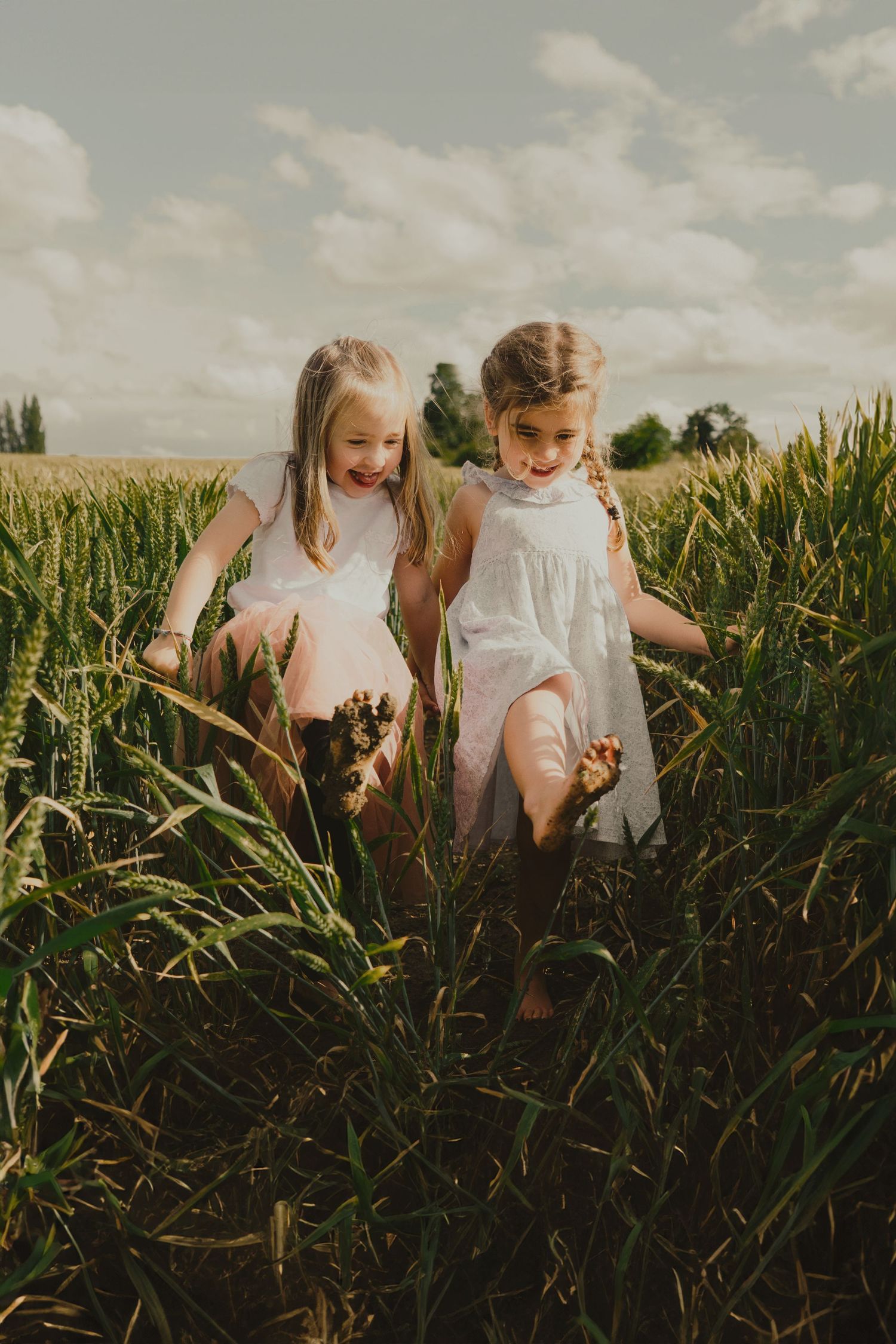 Two girls in white dresses laughing and running barefoot through tall green wheat field on cloudy day.