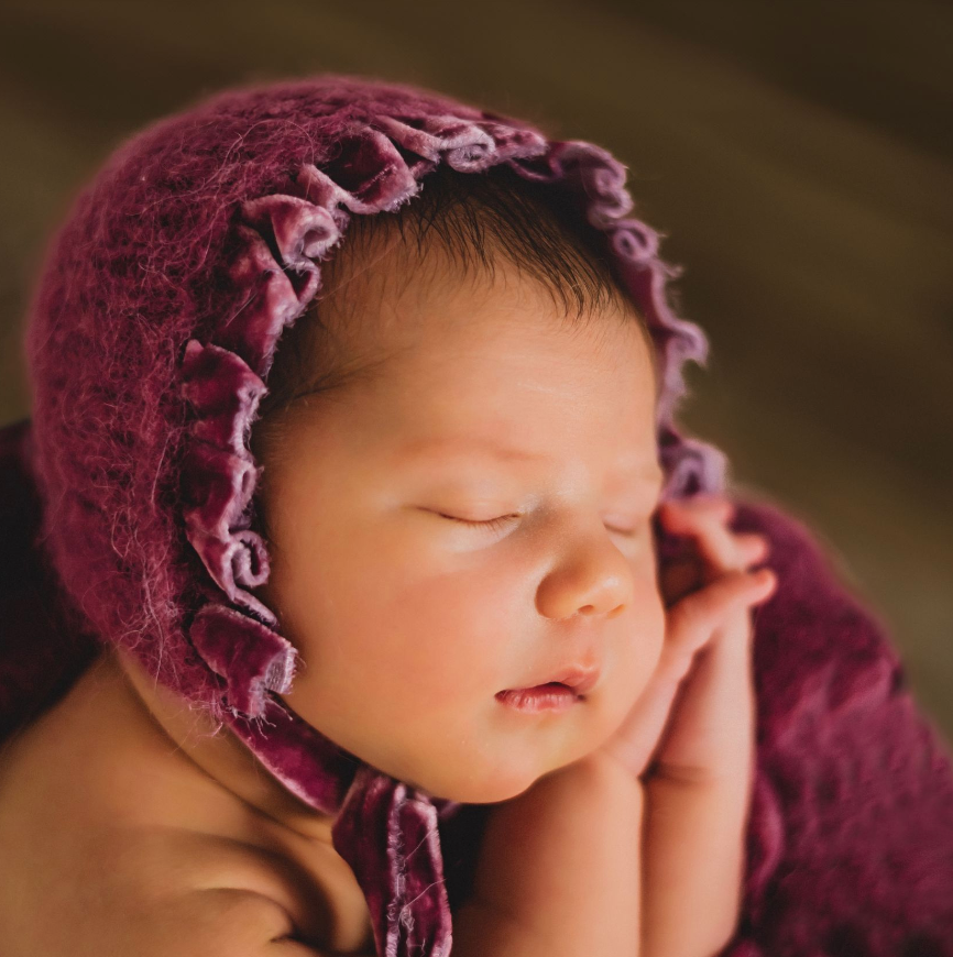 Sleeping newborn baby wearing a purple knit bonnet, wrapped in a soft magenta blanket.