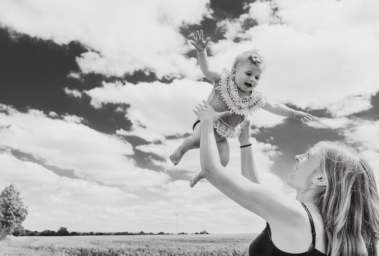 Mother joyfully tosses laughing baby into the air against a dramatic cloudy sky in a field.