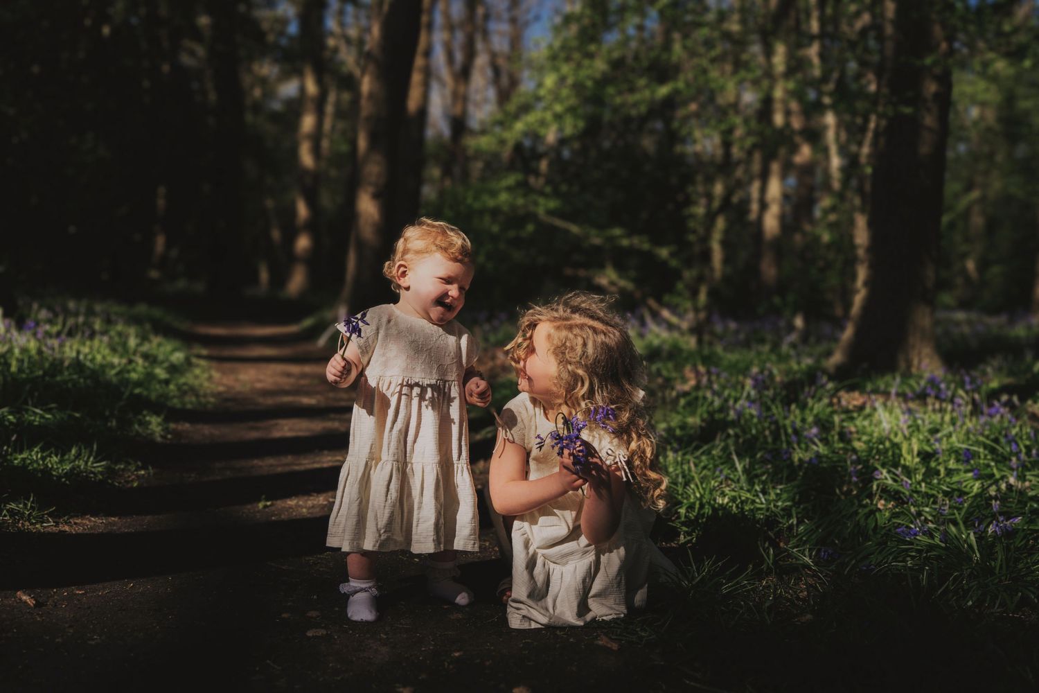Two toddler girls in white dresses share flowers on a sunlit bluebell woodland path.