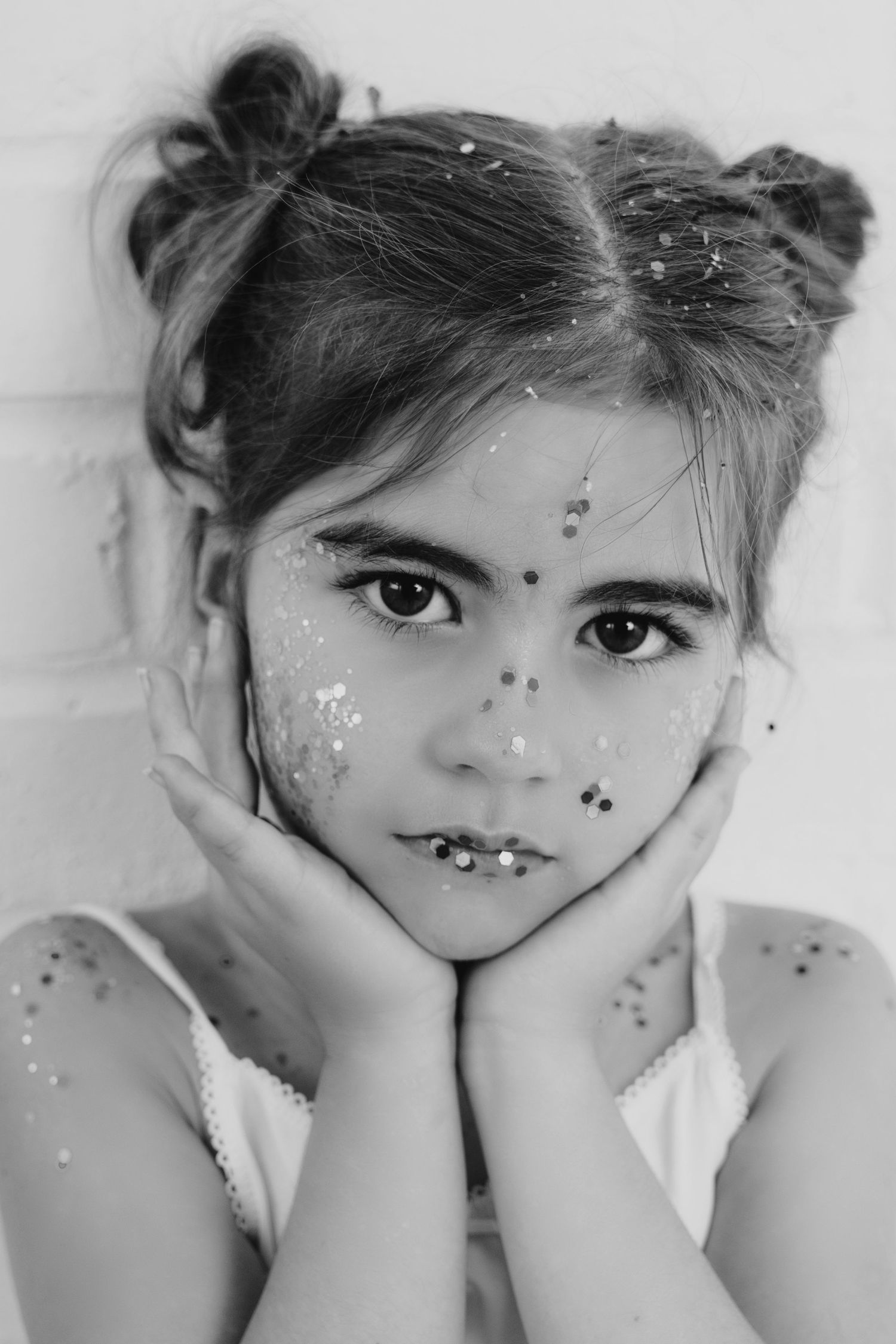 Black and white portrait of a young girl with glitter on her face resting chin in hands.