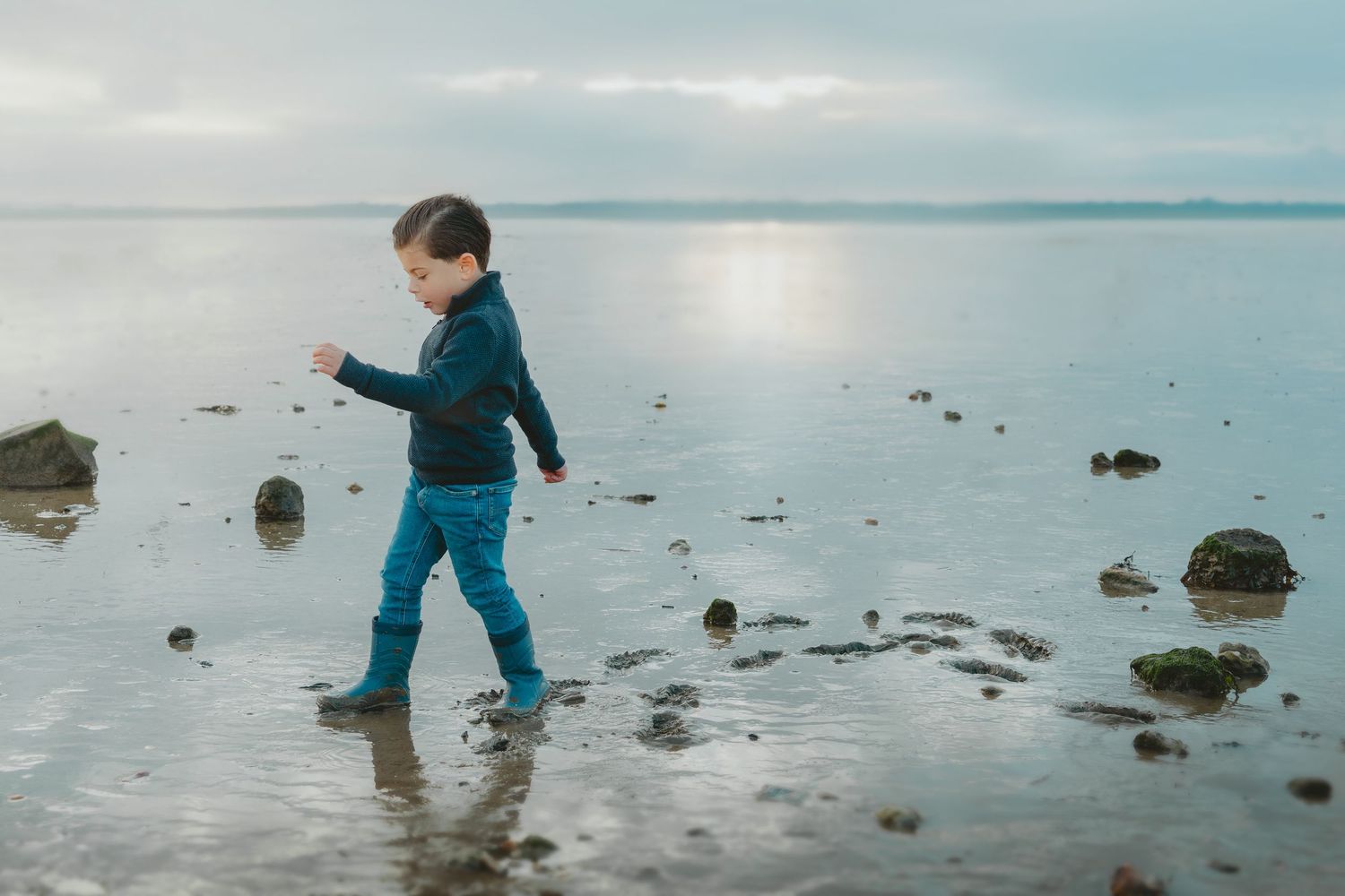 Young boy in blue rain boots walks along a rocky shoreline under a cloudy sky.