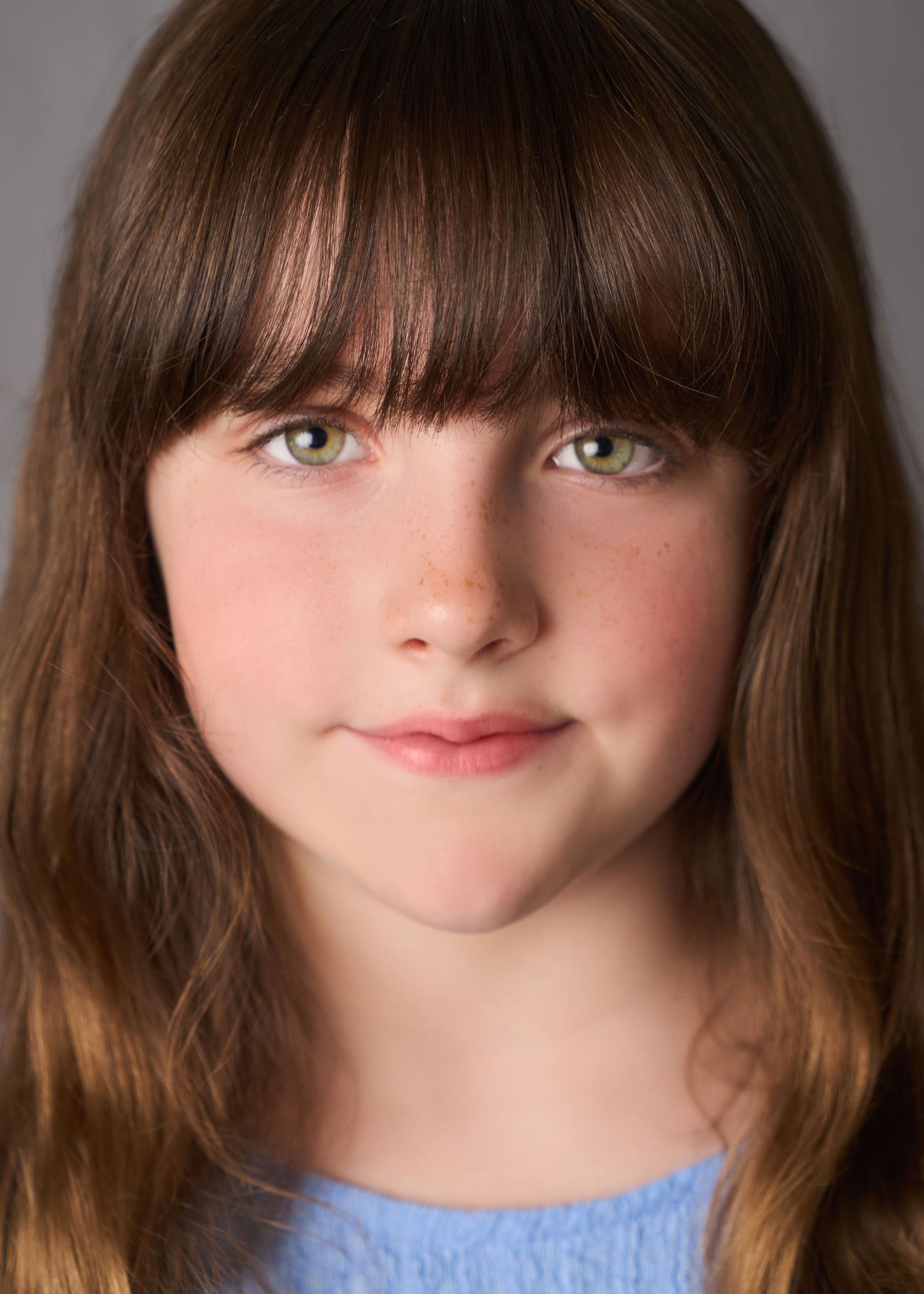 Young girl with green eyes, freckles, and brown bangs smiling softly in a blue top against gray background.