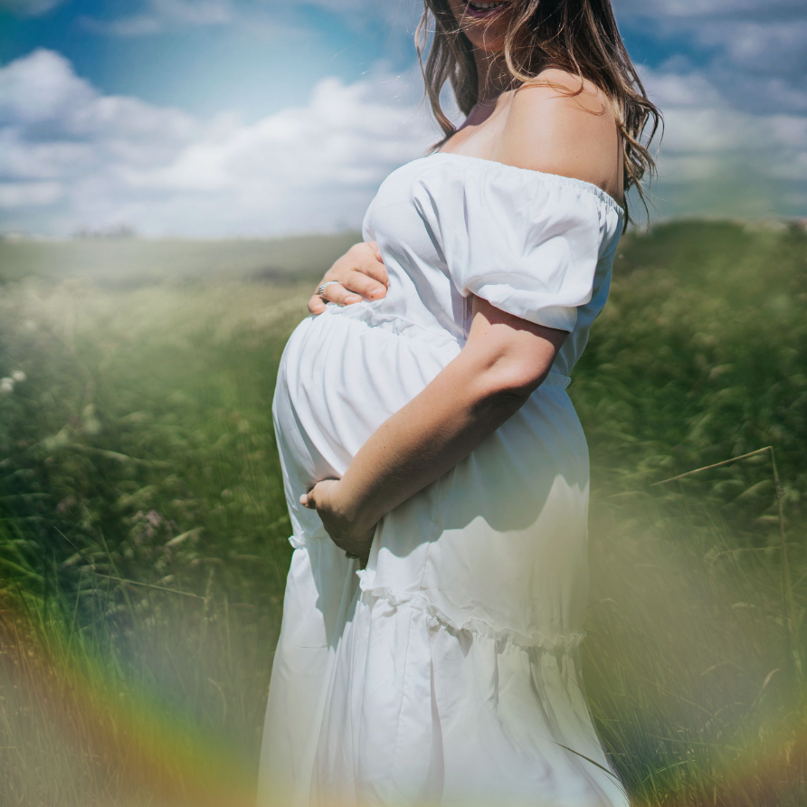 Pregnant woman in white off-shoulder dress cradling her baby bump in a lush green field under a bright blue sky.