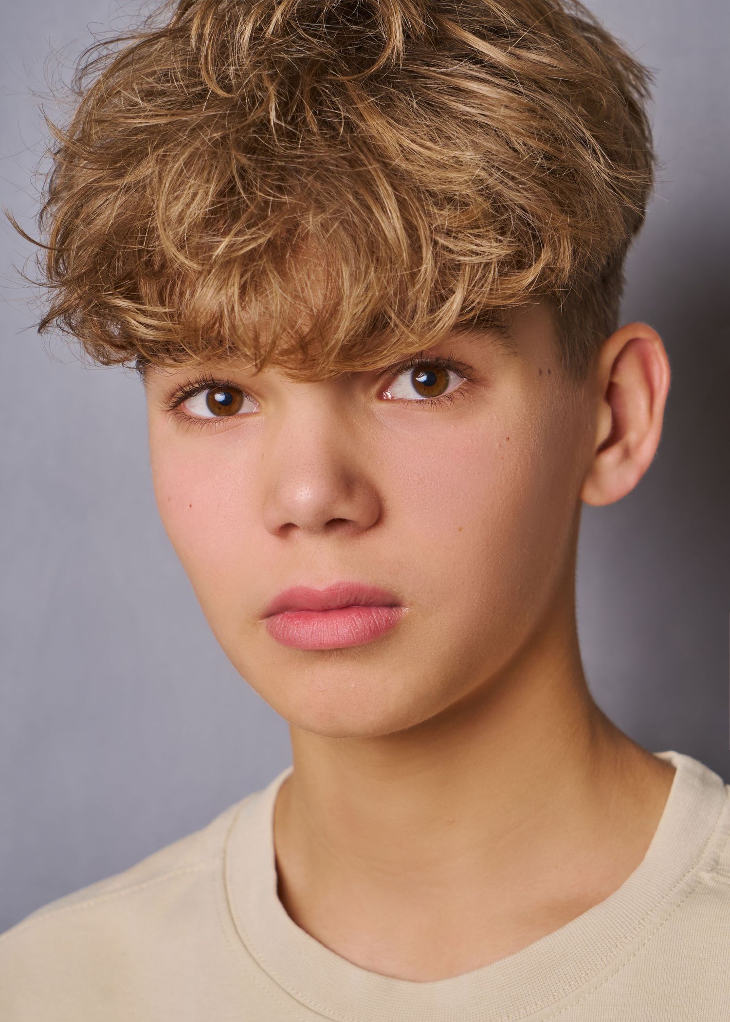 A close up headshot of a teenage boy with short blonde  hair that is medium length and curly on top.