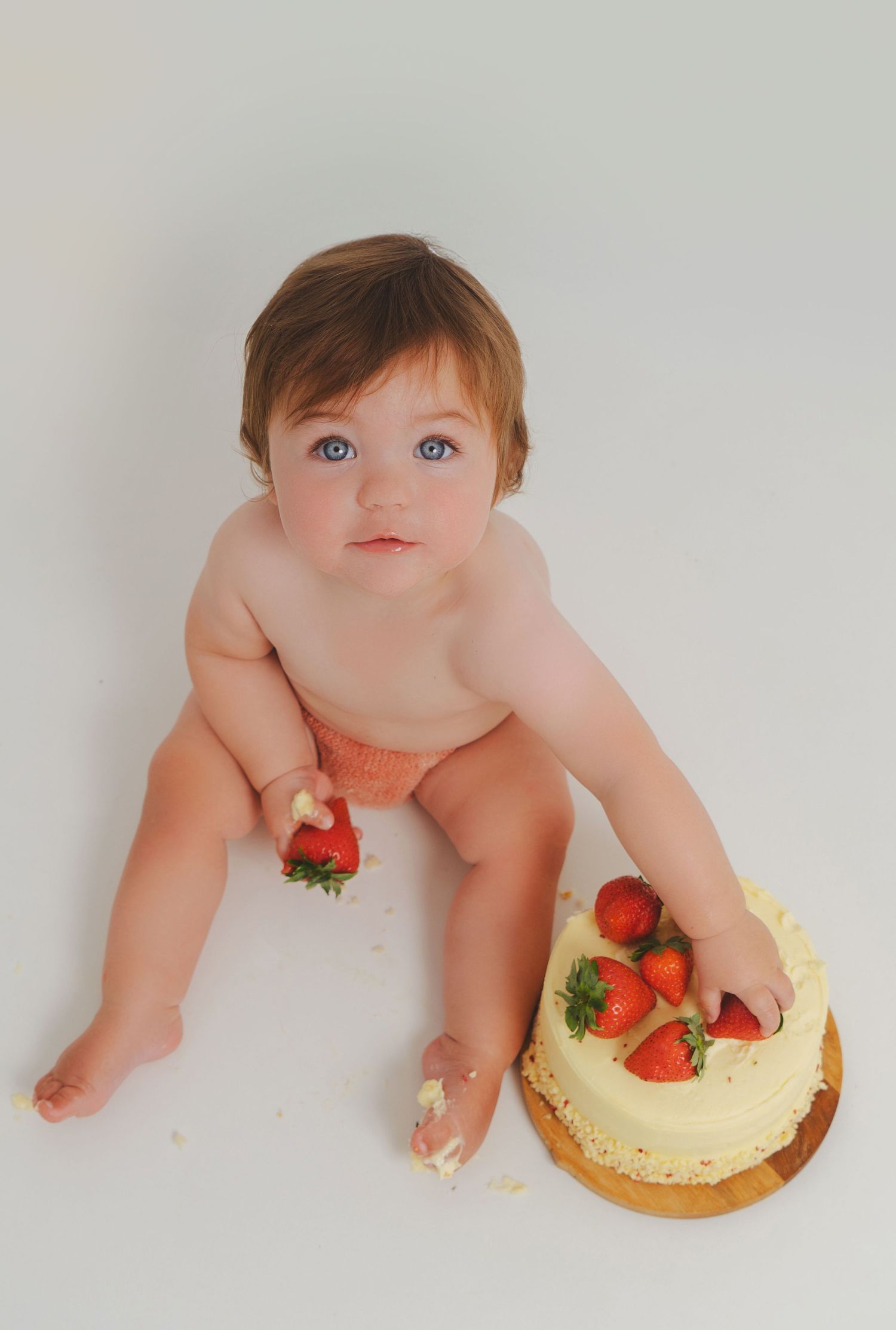 Blue-eyed baby in diaper sitting on white background with a strawberry cake during a cake smash session.