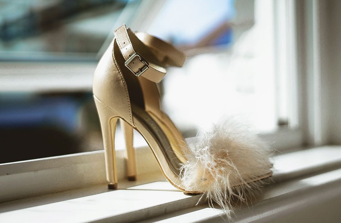 Elegant gold stiletto wedding heel with feather detail displayed on a windowsill.