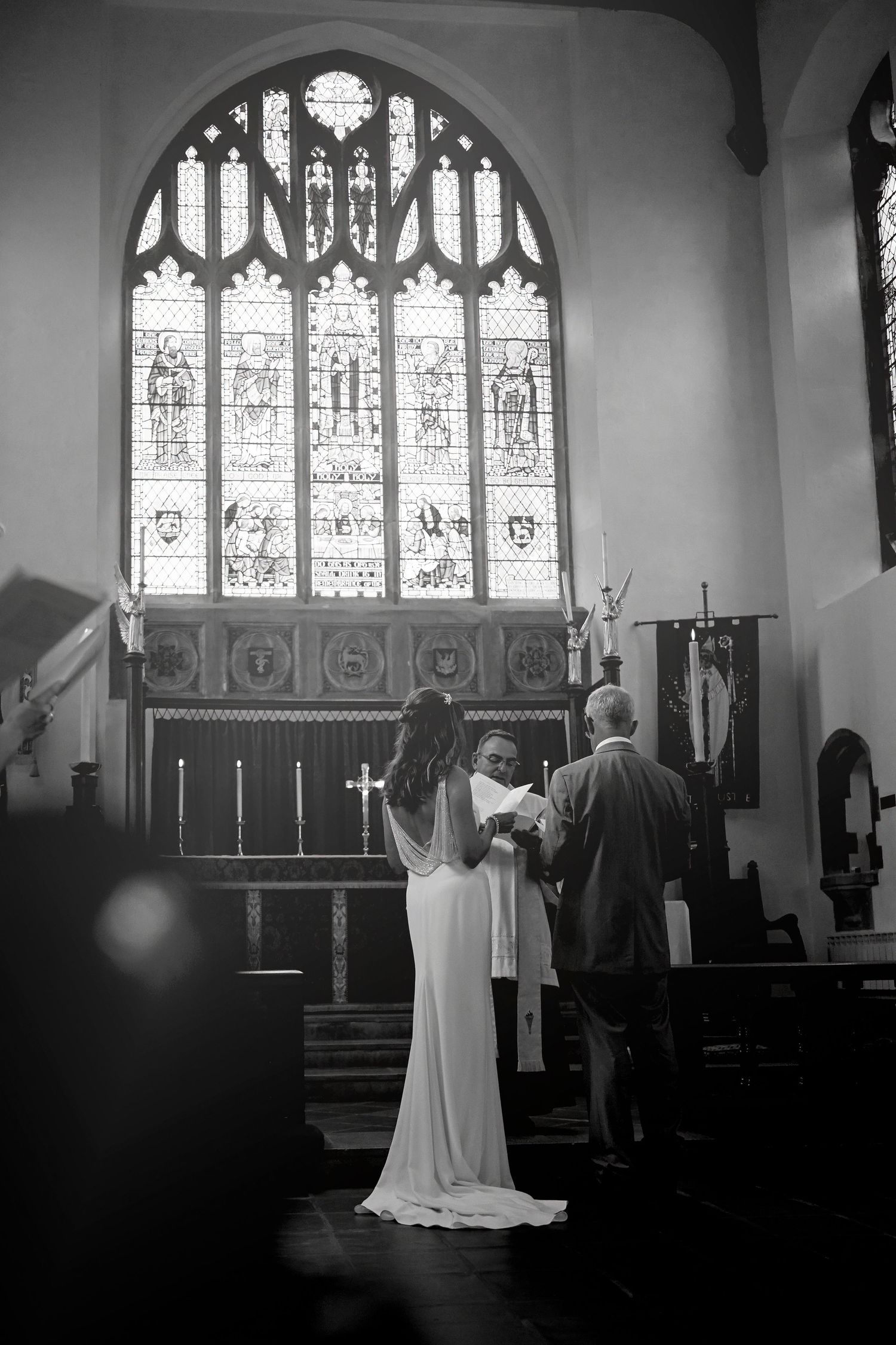 Black and white image of a bride and officiant at a church altar with stunning stained glass.