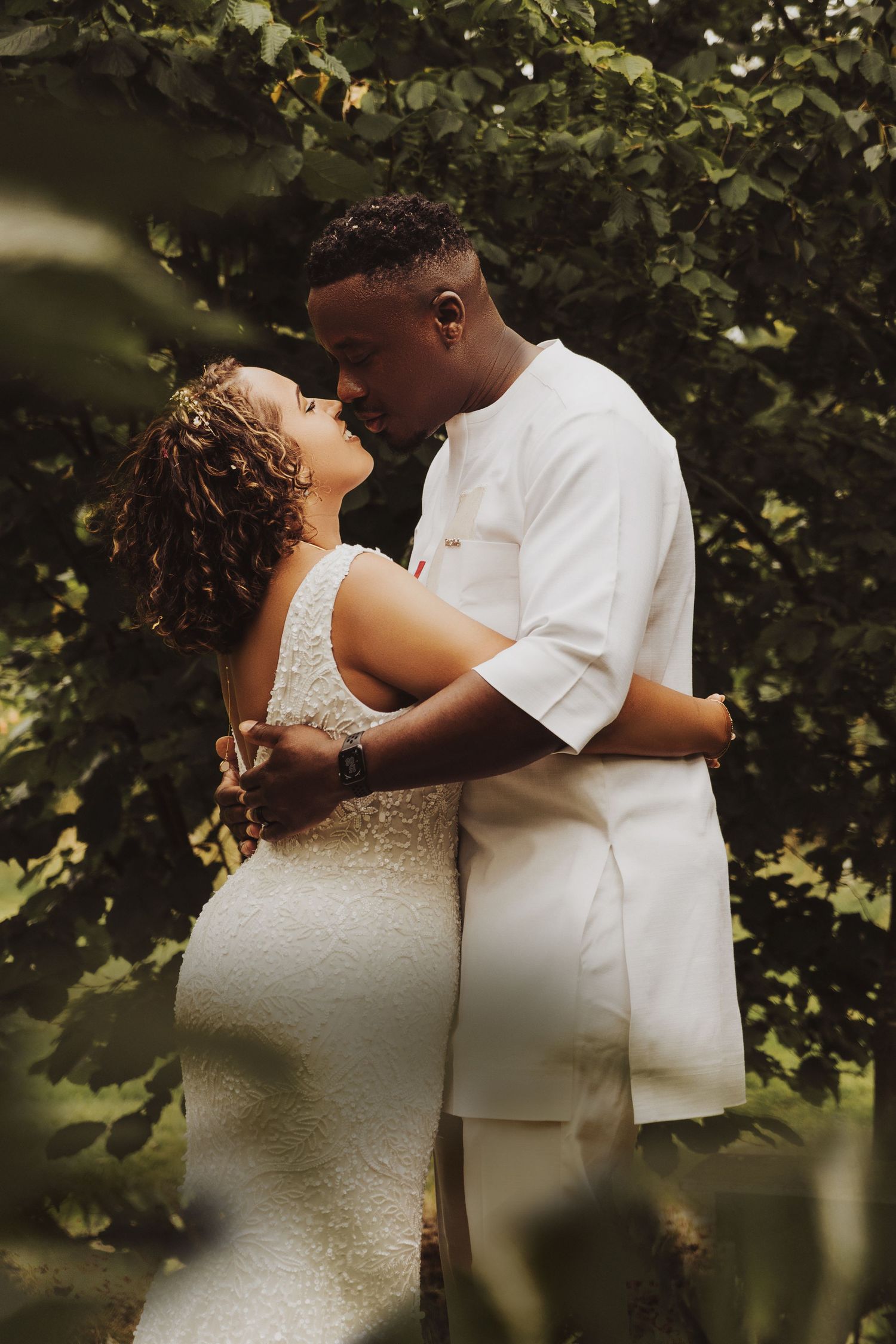 Couple in wedding attire sharing intimate embrace surrounded by lush green foliage in garden setting.