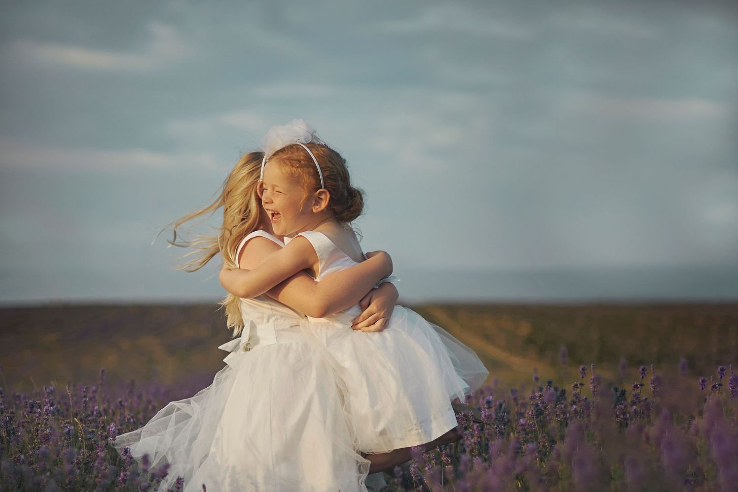 Two little girls in white dresses hugging in a lavender field under a cloudy sky.