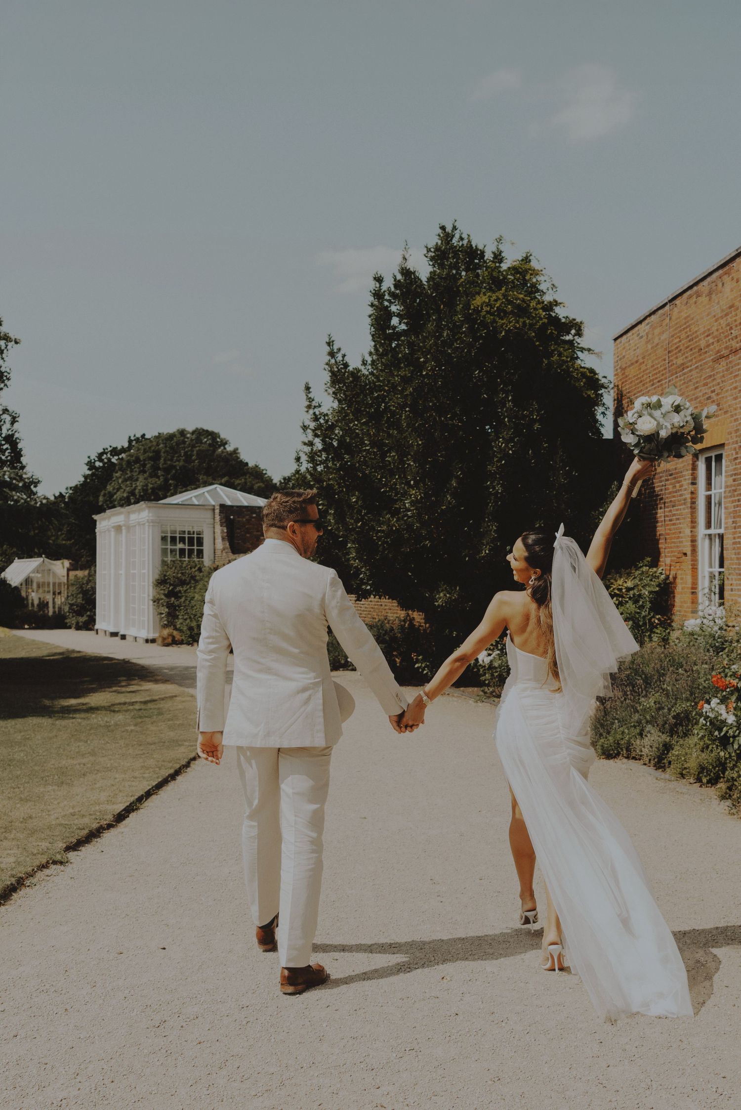 Bride raising bouquet joyfully while walking hand-in-hand with groom along a sunny garden path.
