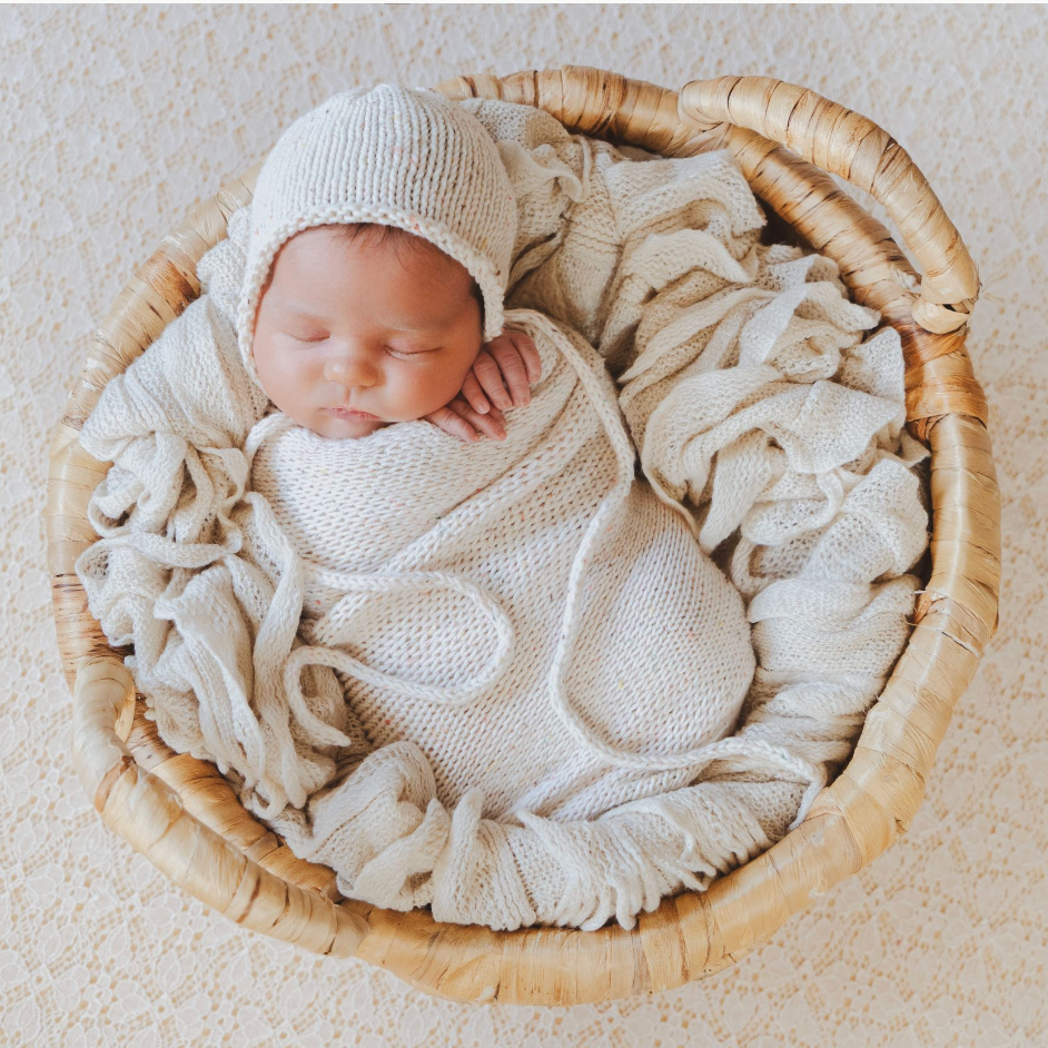 Sleeping newborn baby wrapped in cream knit swaddle and bonnet, nestled in a wicker basket with soft fabric.