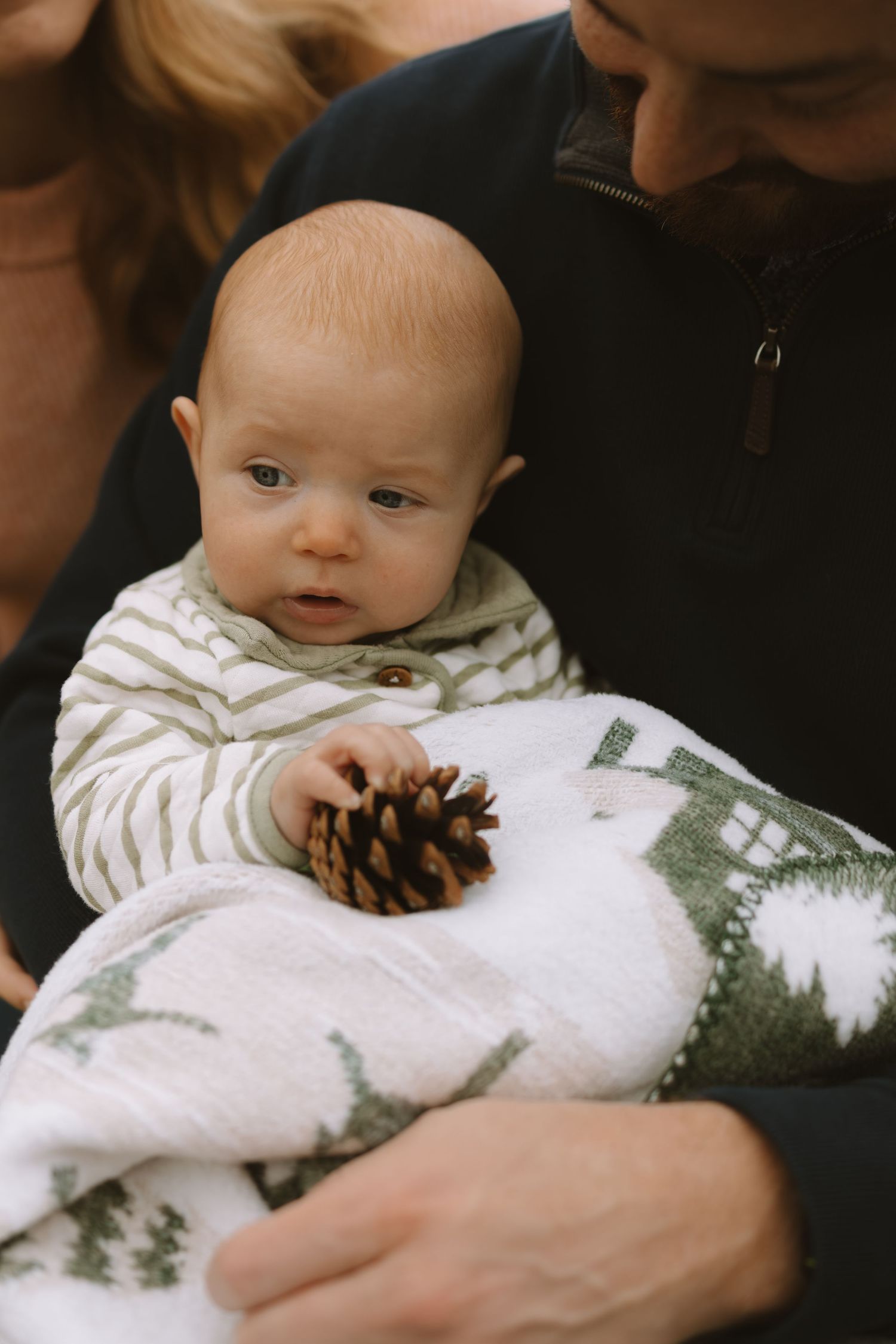 Baby wrapped in white pine tree print blanket holds pinecone during photo session.