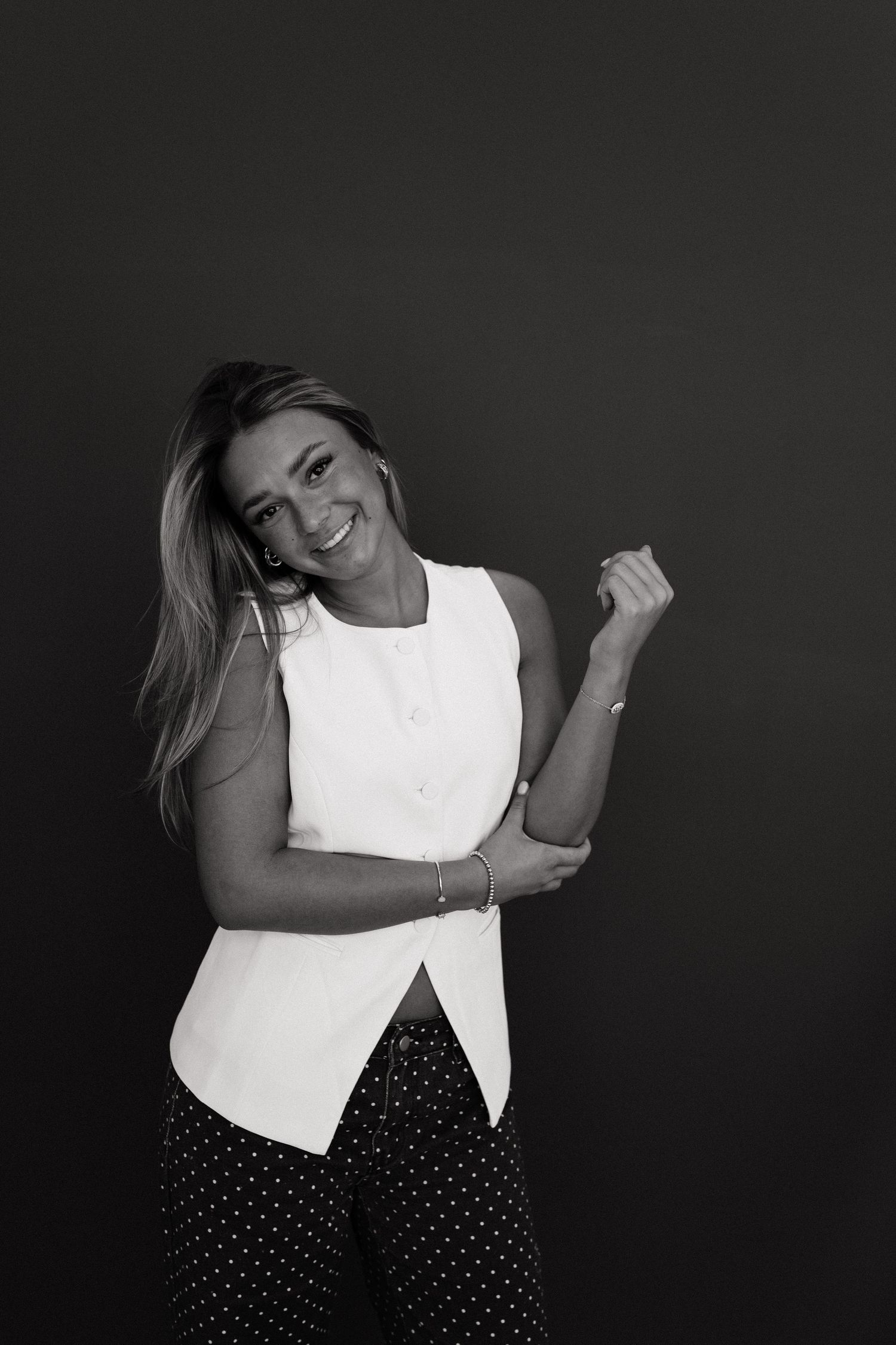 A person in a white sleeveless top and polka dot skirt poses playfully against a dark background.