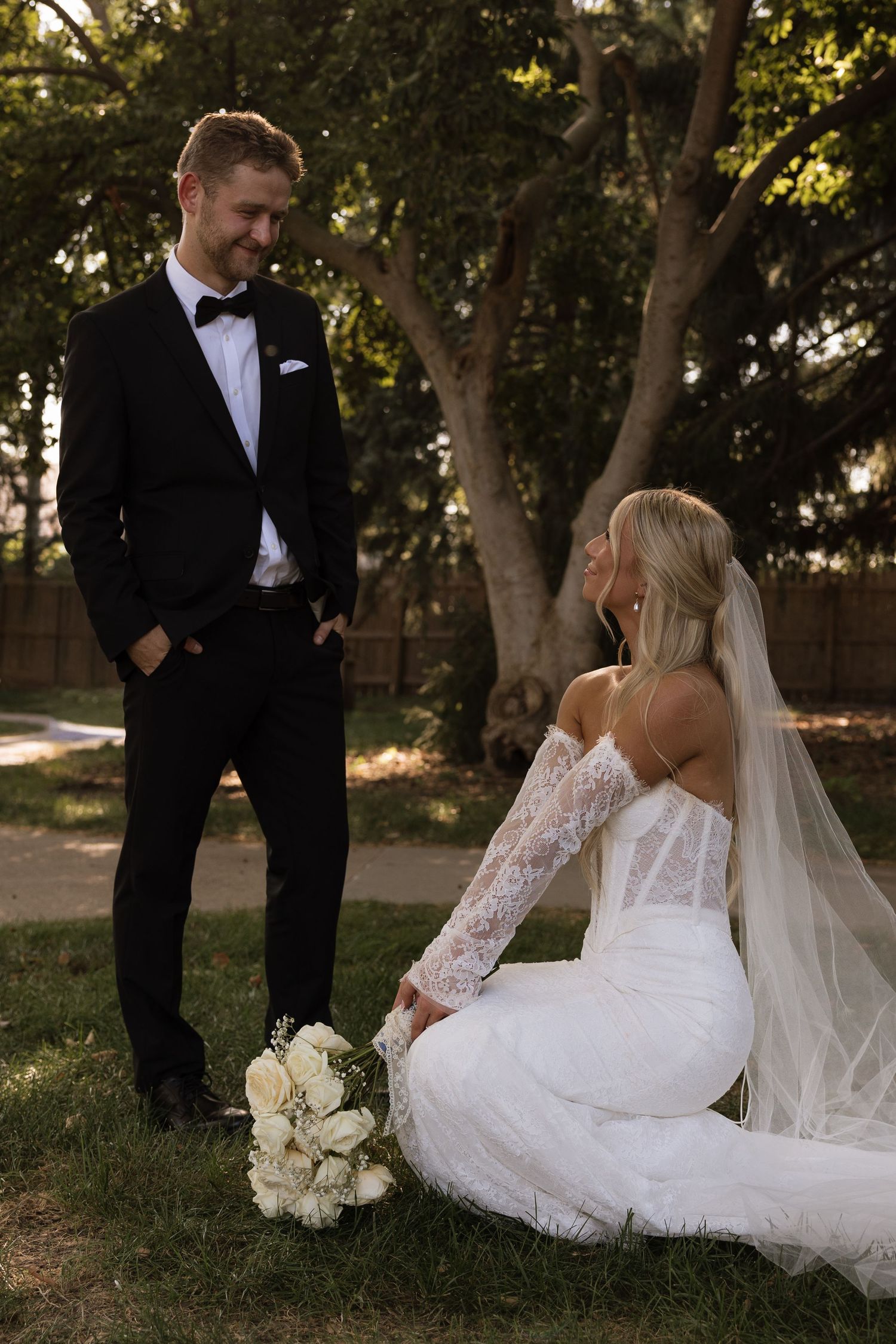 Bride in lace off-shoulder gown sits on grass holding white bouquet as groom in black tuxedo smiles outdoors.