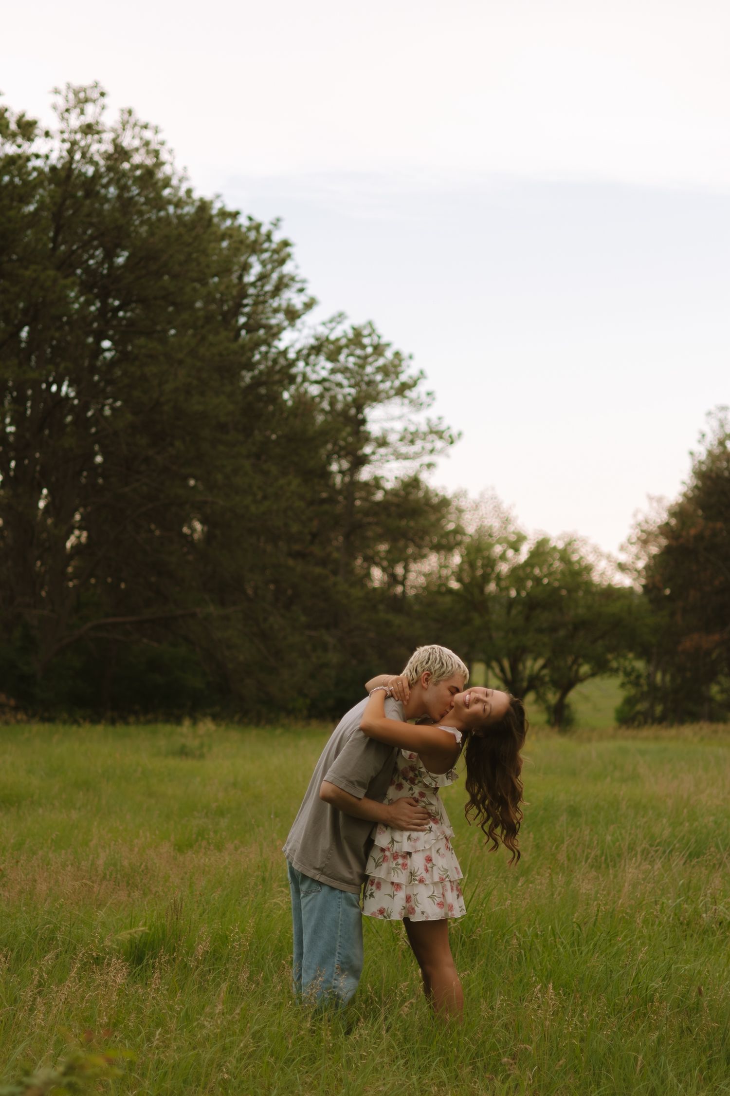A romantic couple embraces in a sunlit meadow surrounded by trees during golden hour.
