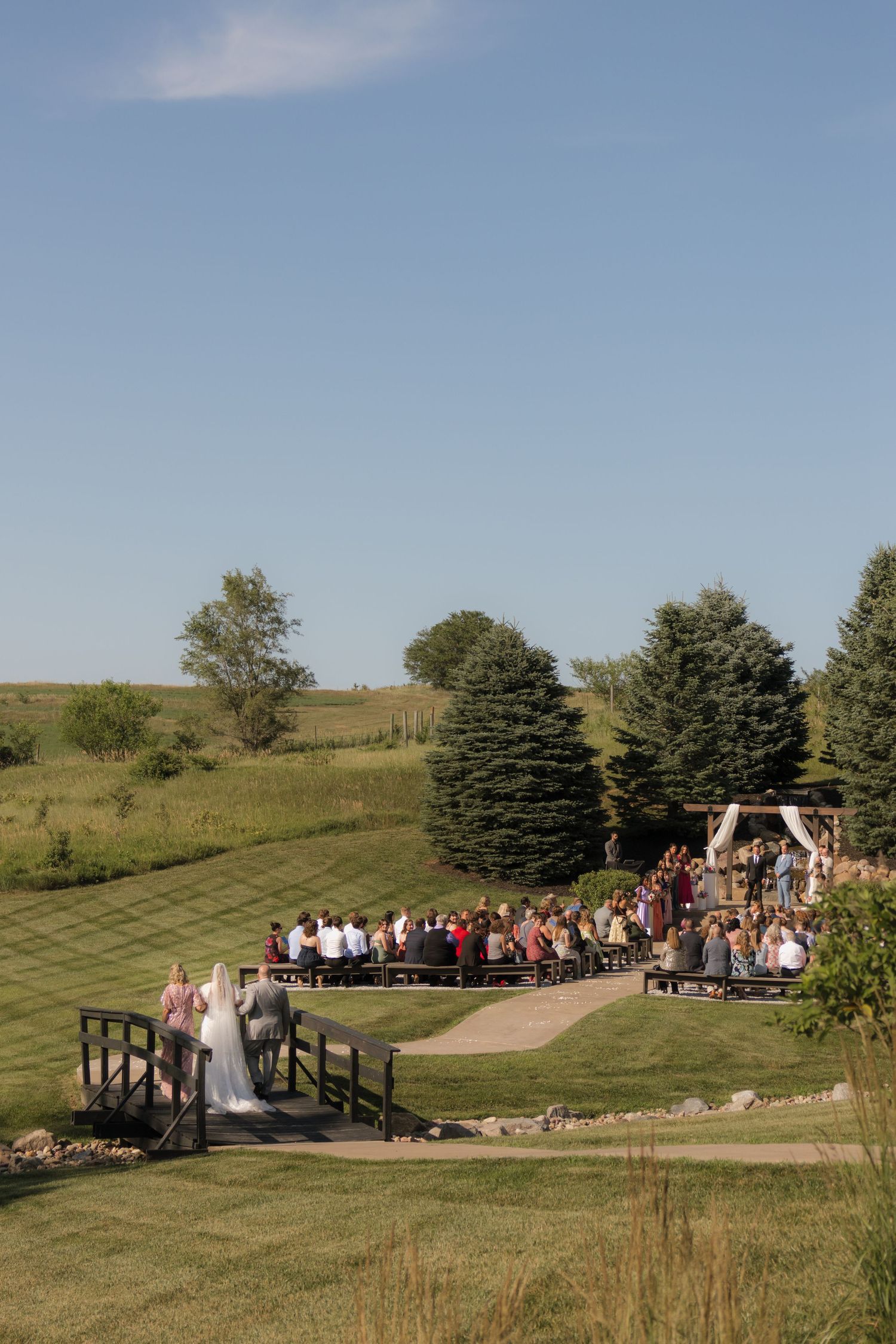 Outdoor wedding ceremony setup on rolling hills with guests gathered on wooden bridge.
