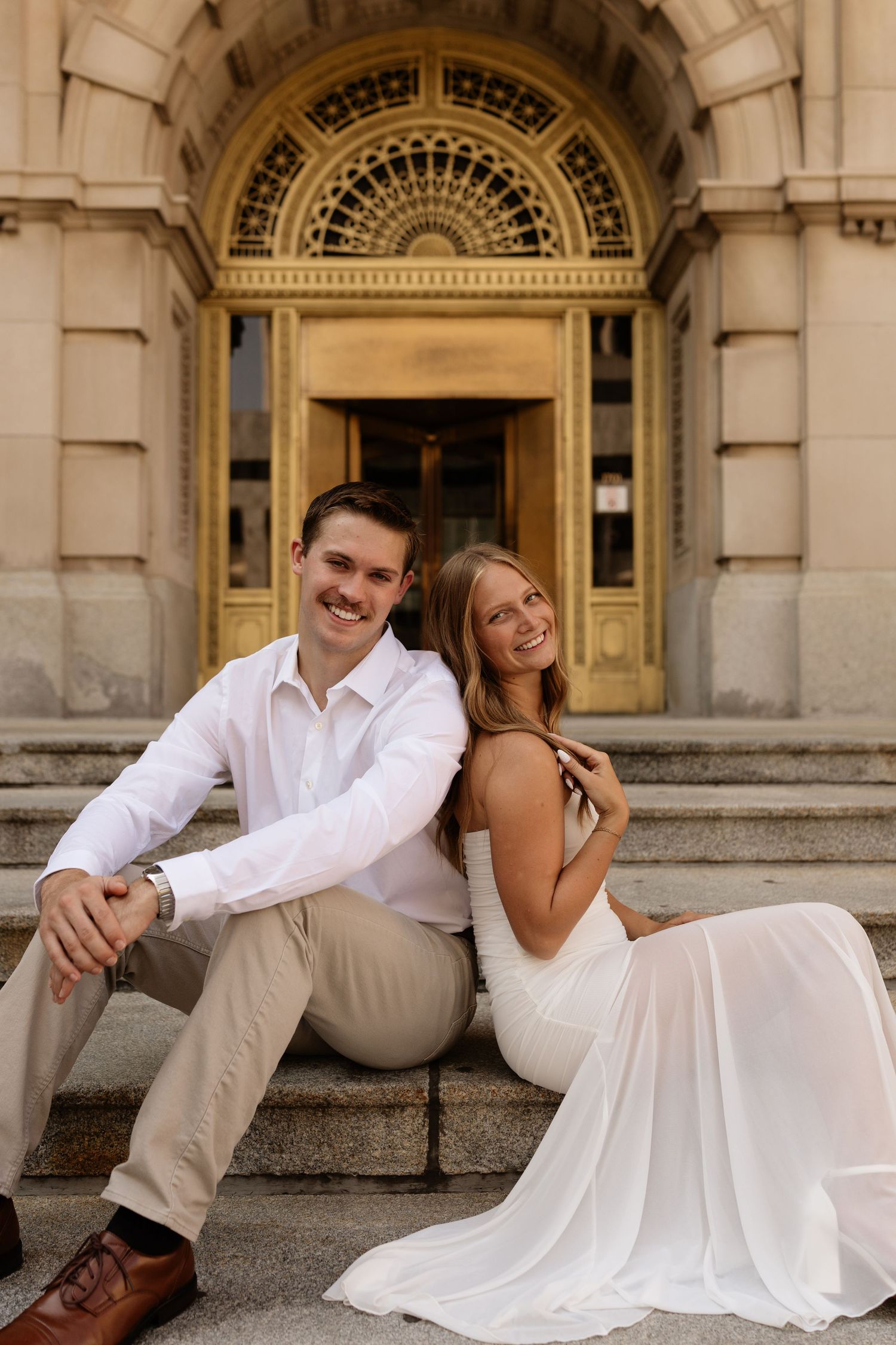 A smiling couple in wedding attire sits on ornate building steps, woman in white gown, man in white shirt and tan pants.