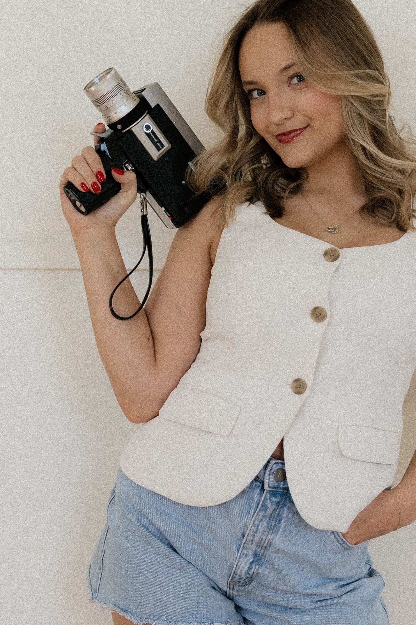 A person wearing a white sleeveless top and light blue jeans poses with a vintage camera against a neutral background.