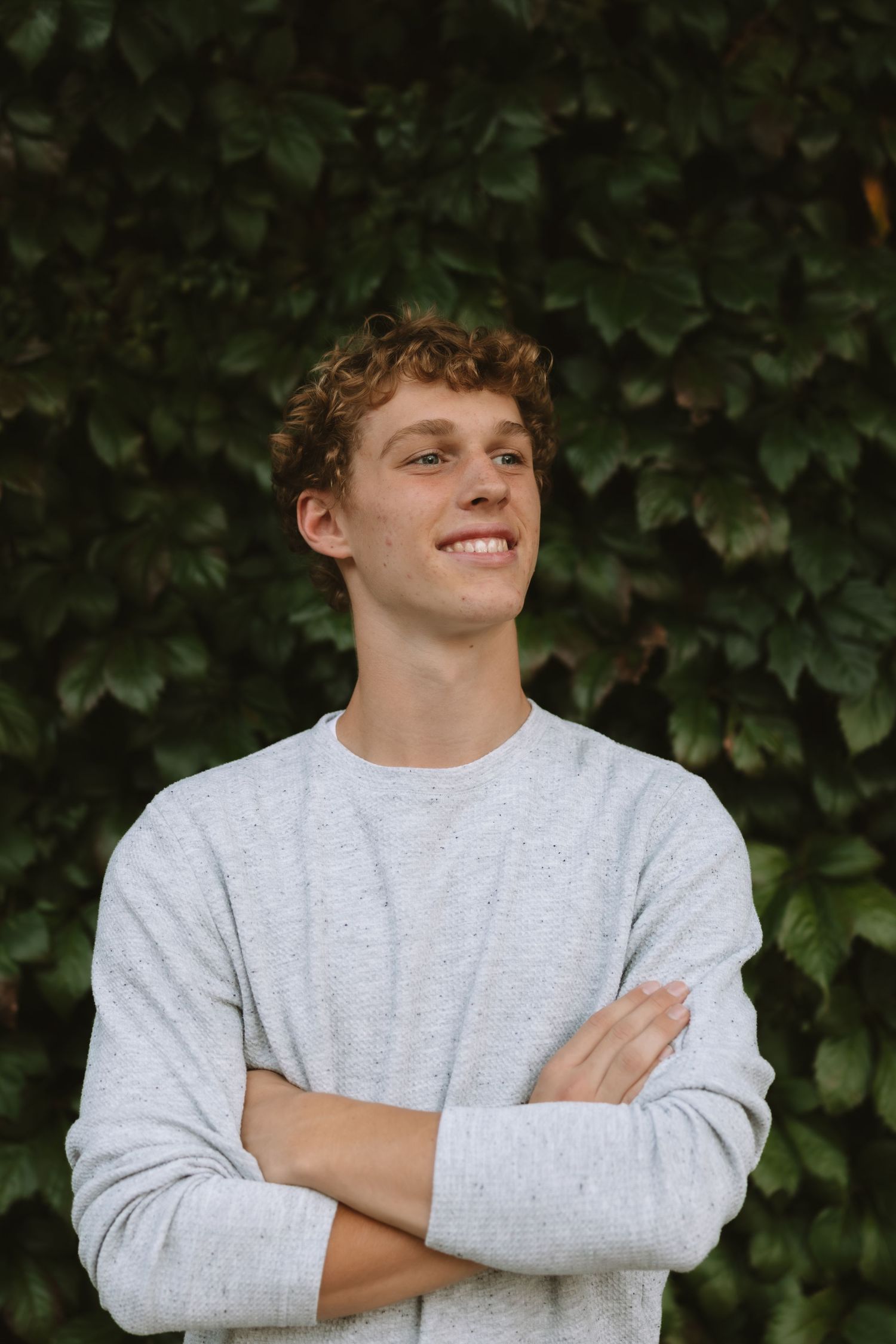 Student in light gray sweatshirt smiles against green leafy backdrop.