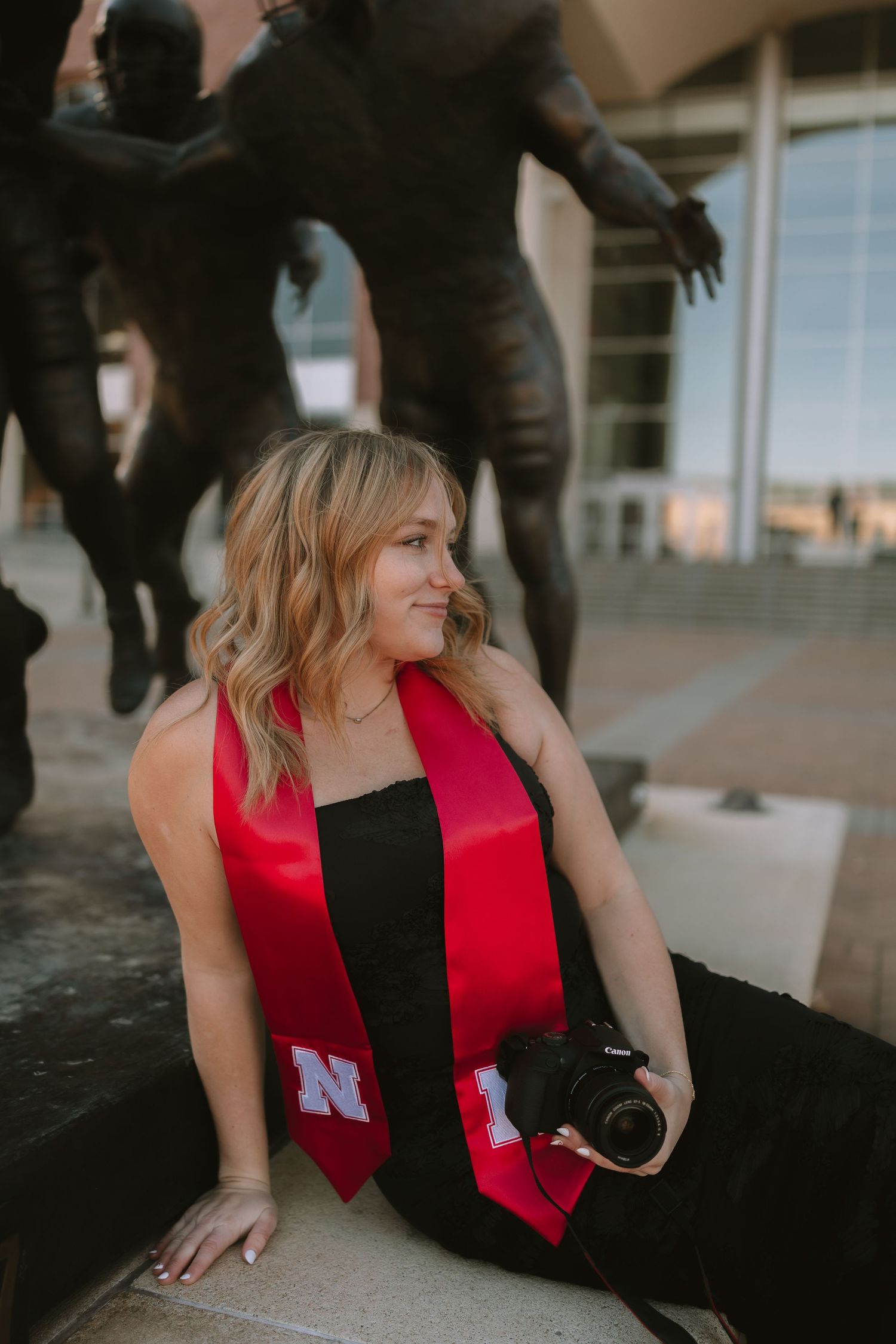 Graduate in red and black graduation stole poses by statue outside building.
