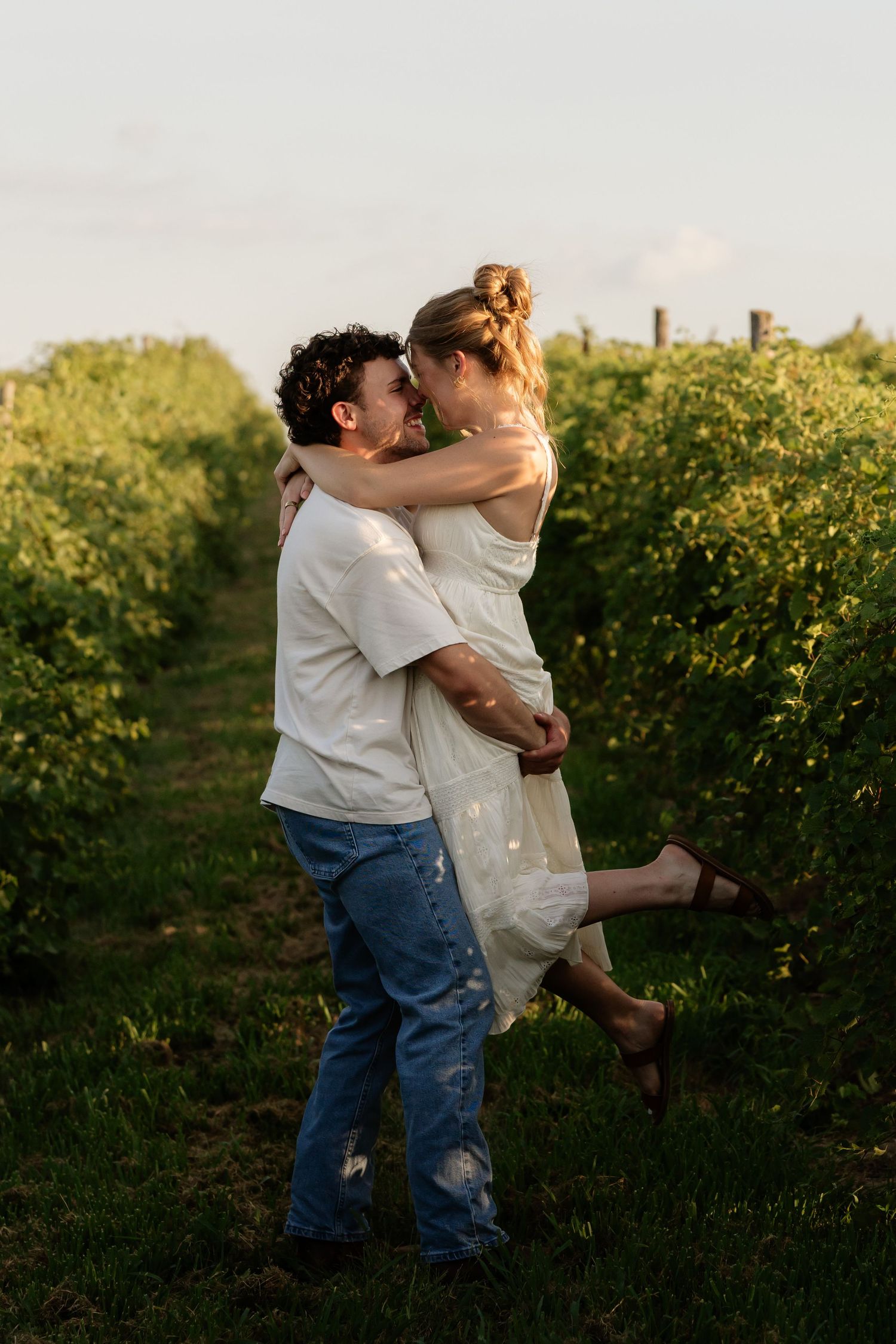 Couple sharing a romantic embrace in a lush vineyard at golden hour, man lifting woman in white dress.