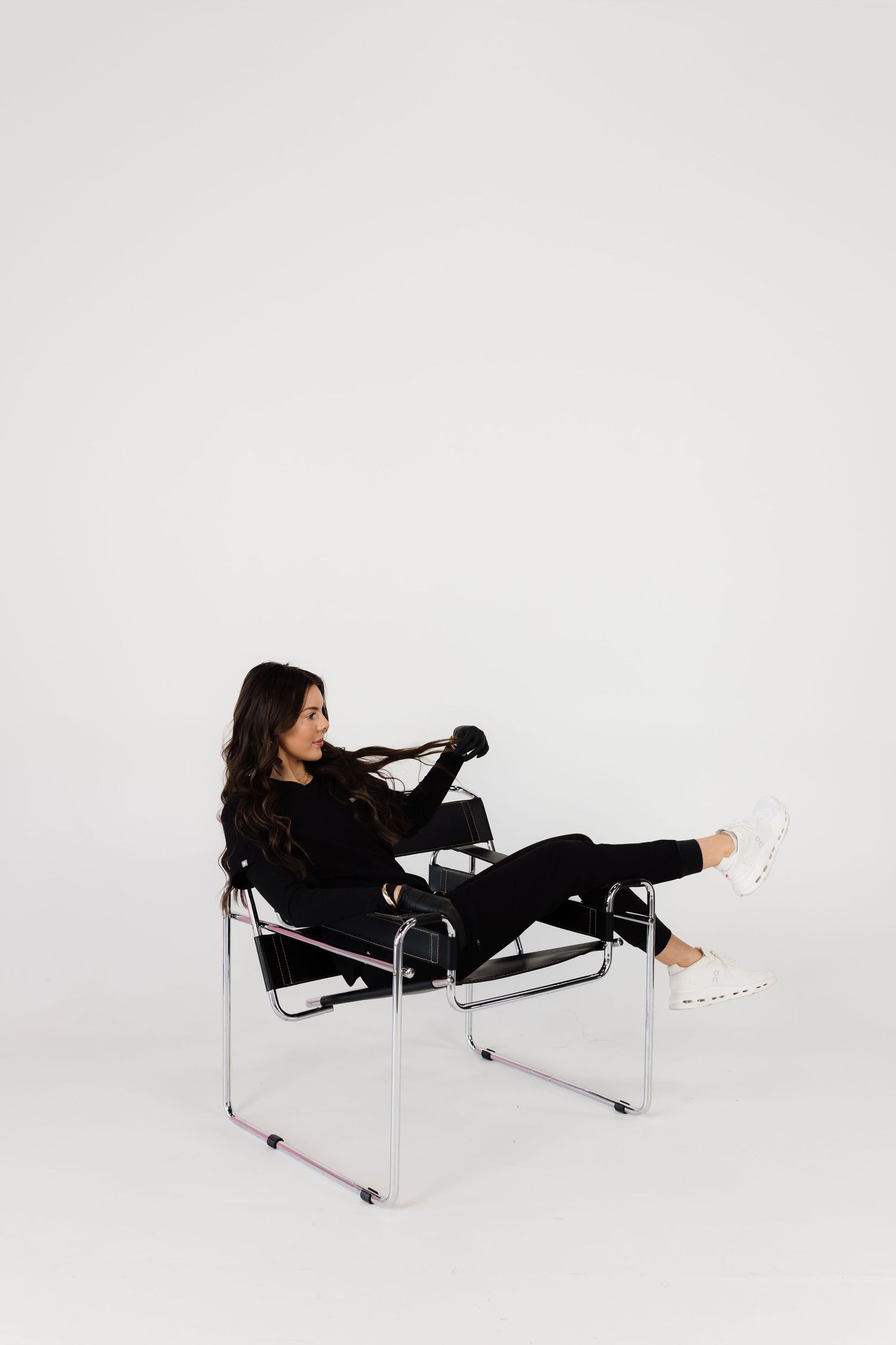 Person in black outfit lounging stylishly on modern chrome chair against white background.