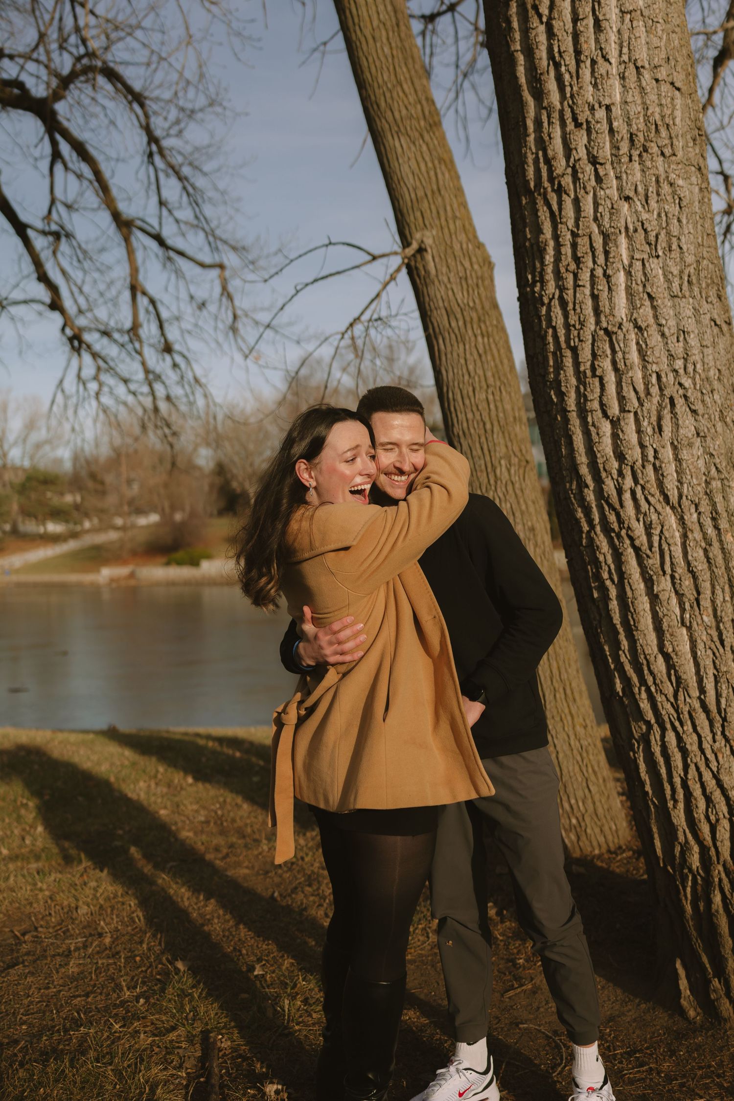 A joyful couple embraces and laughs together near trees by a lakeside.