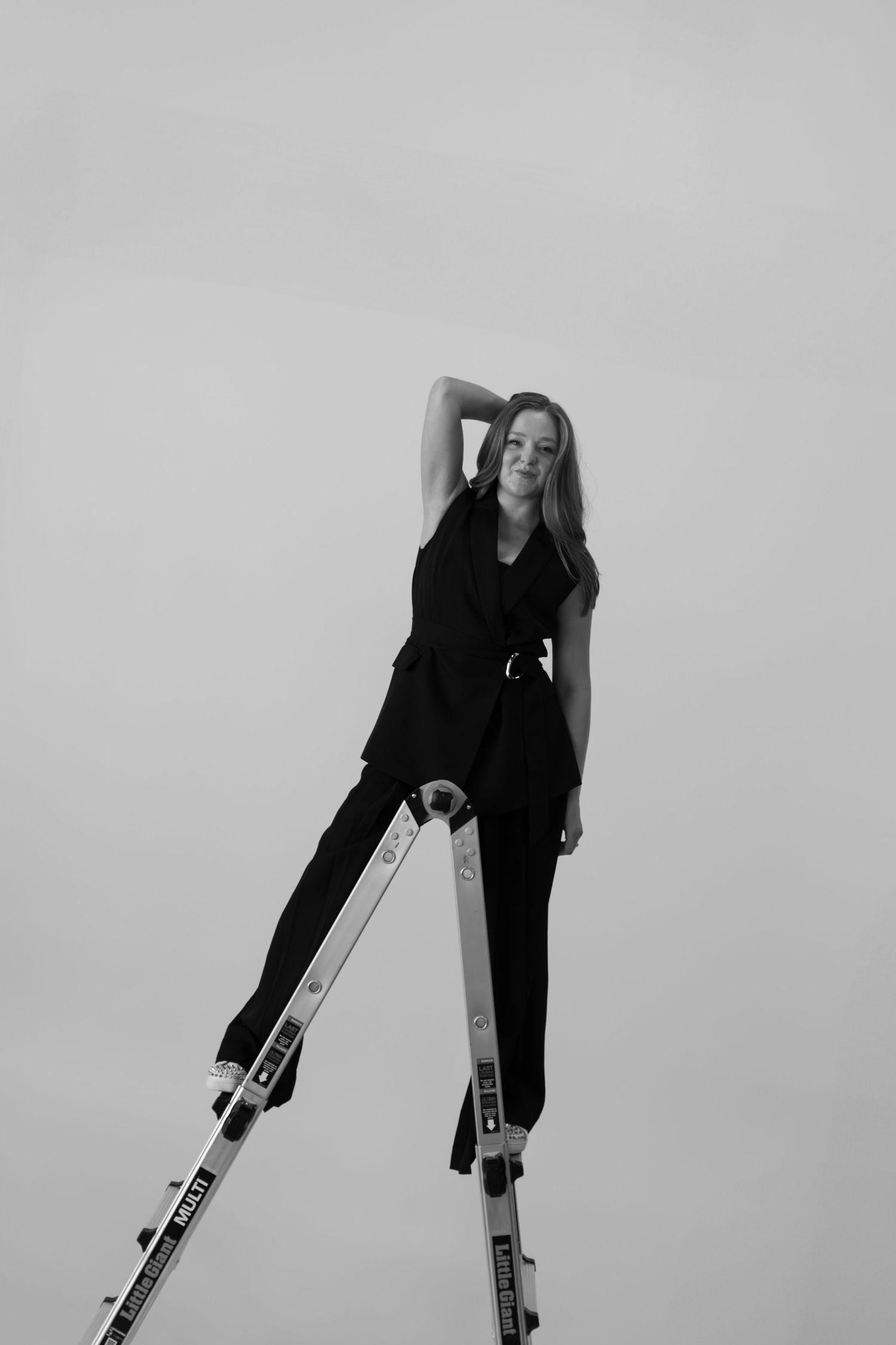 Person in black outfit posing on ladder against minimalist background in monochrome.