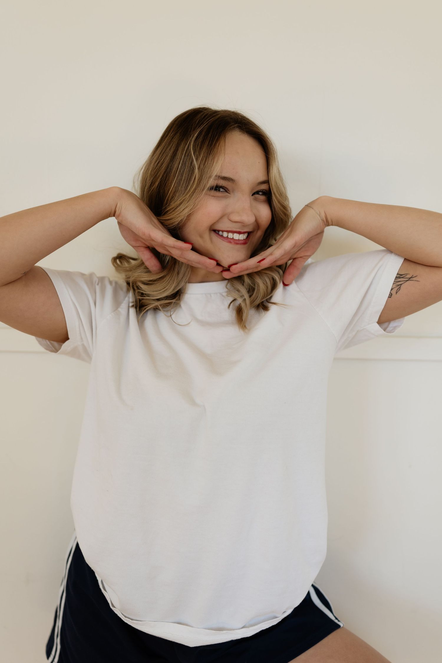 A person in a white t-shirt and dark shorts poses playfully with hands framing their smiling face against a neutral wall.