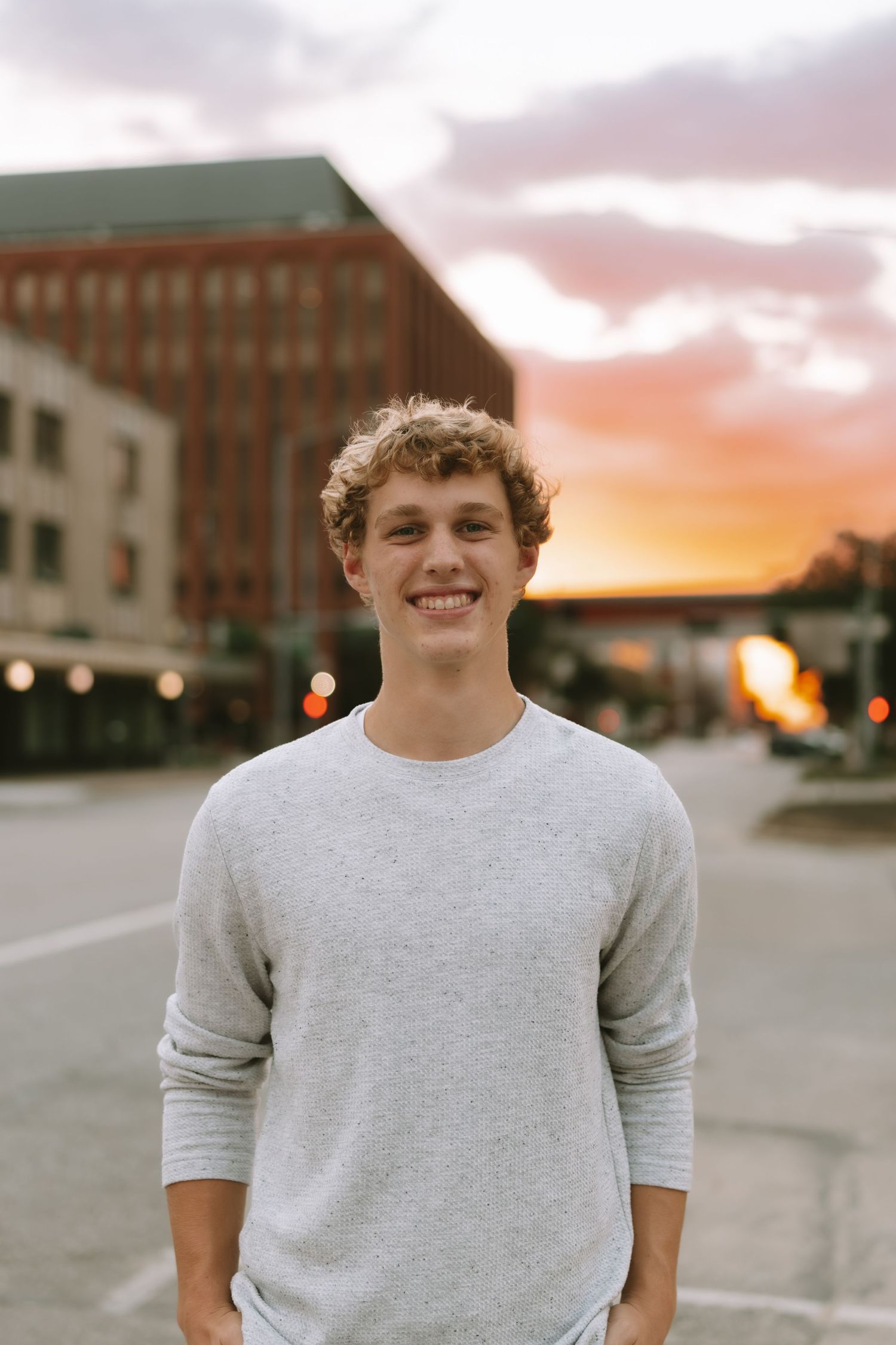 A person in a gray sweater stands on a city street at sunset with buildings in the background.