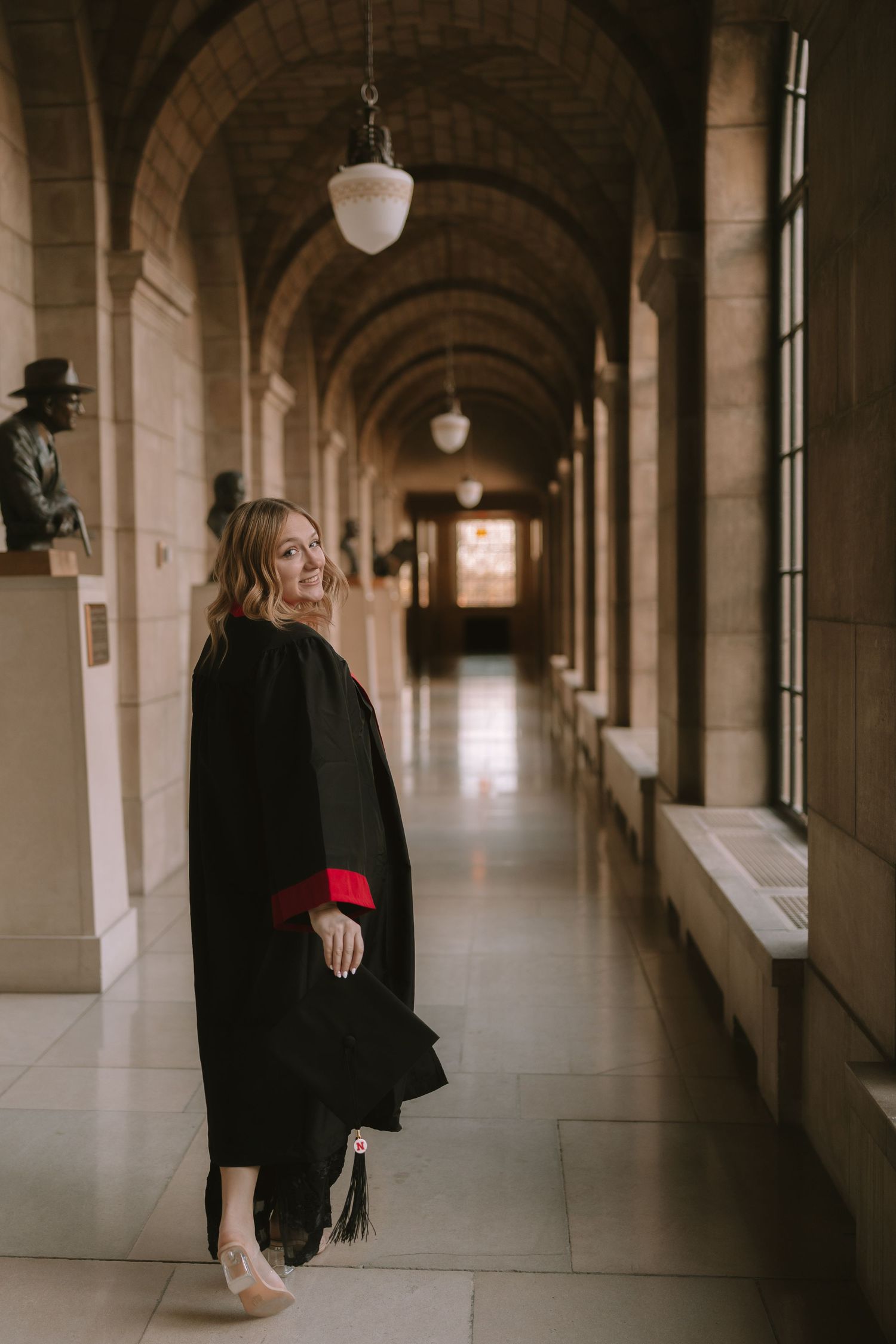Graduate in black cap and gown poses in arched hallway with classic architecture.