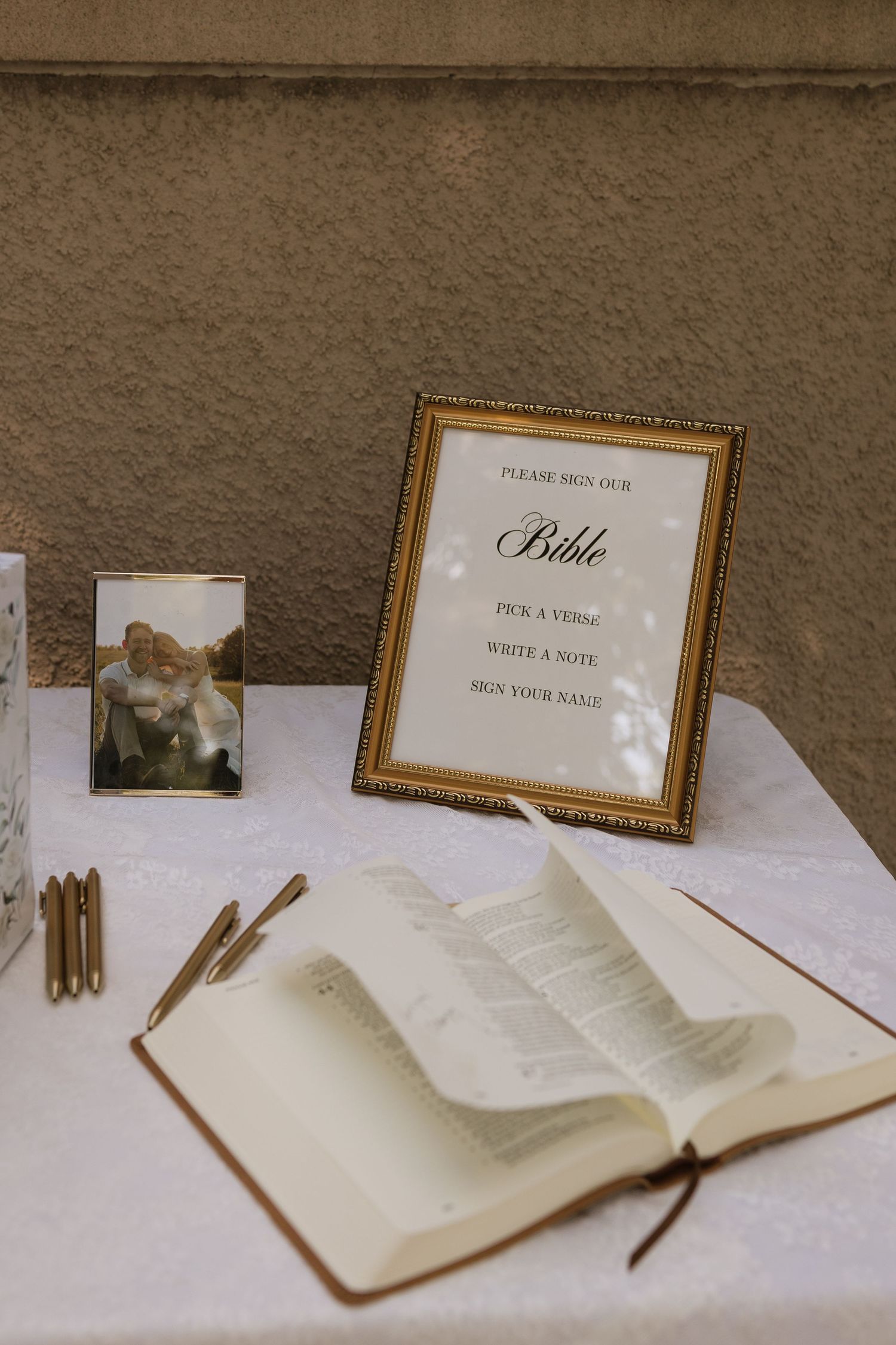 Wedding Bible guest book display with gold frame sign, open Bible, gold pens, and couple photo on white linen table.