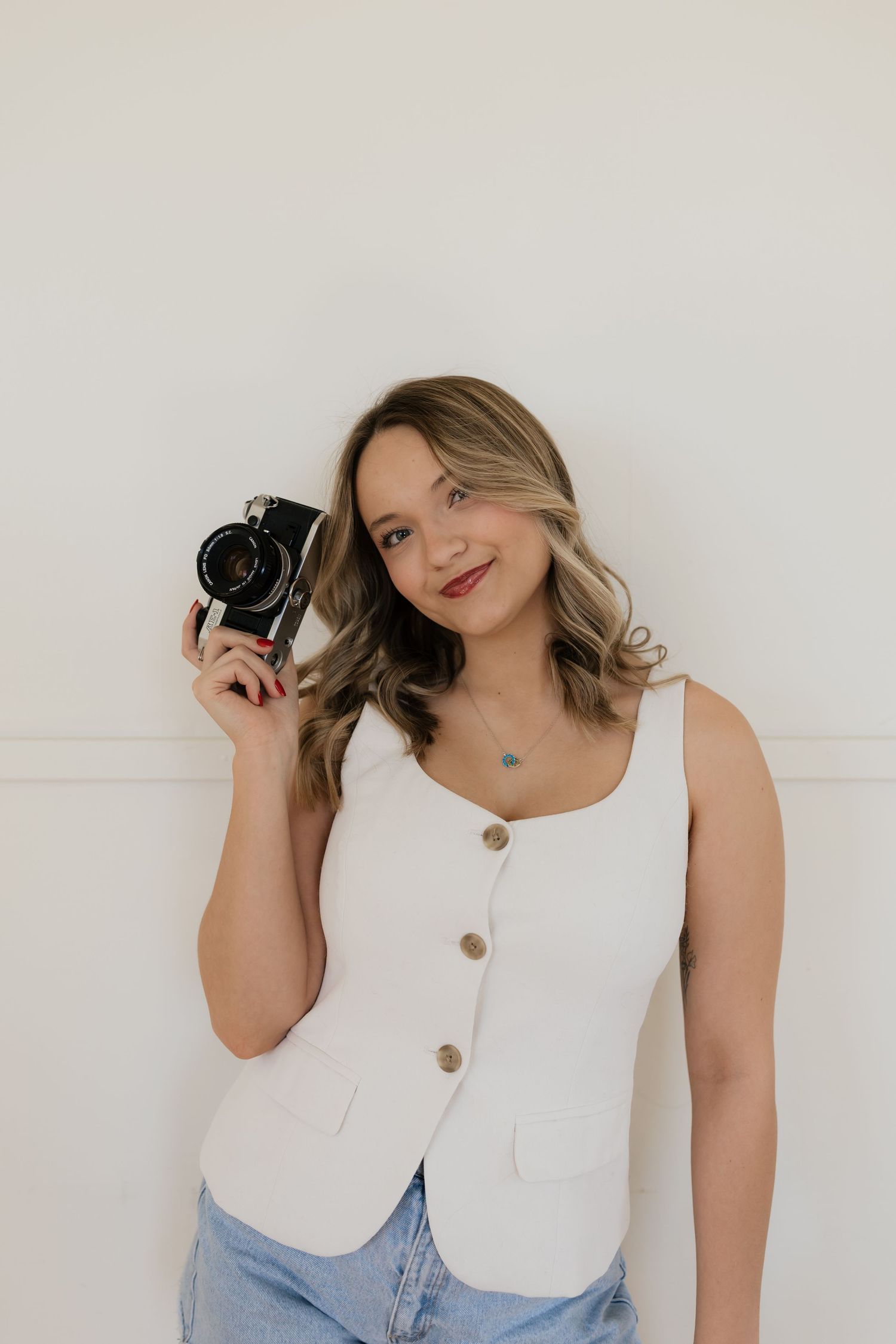 A photographer in a white tank top and jeans poses with a vintage camera against a white wall.