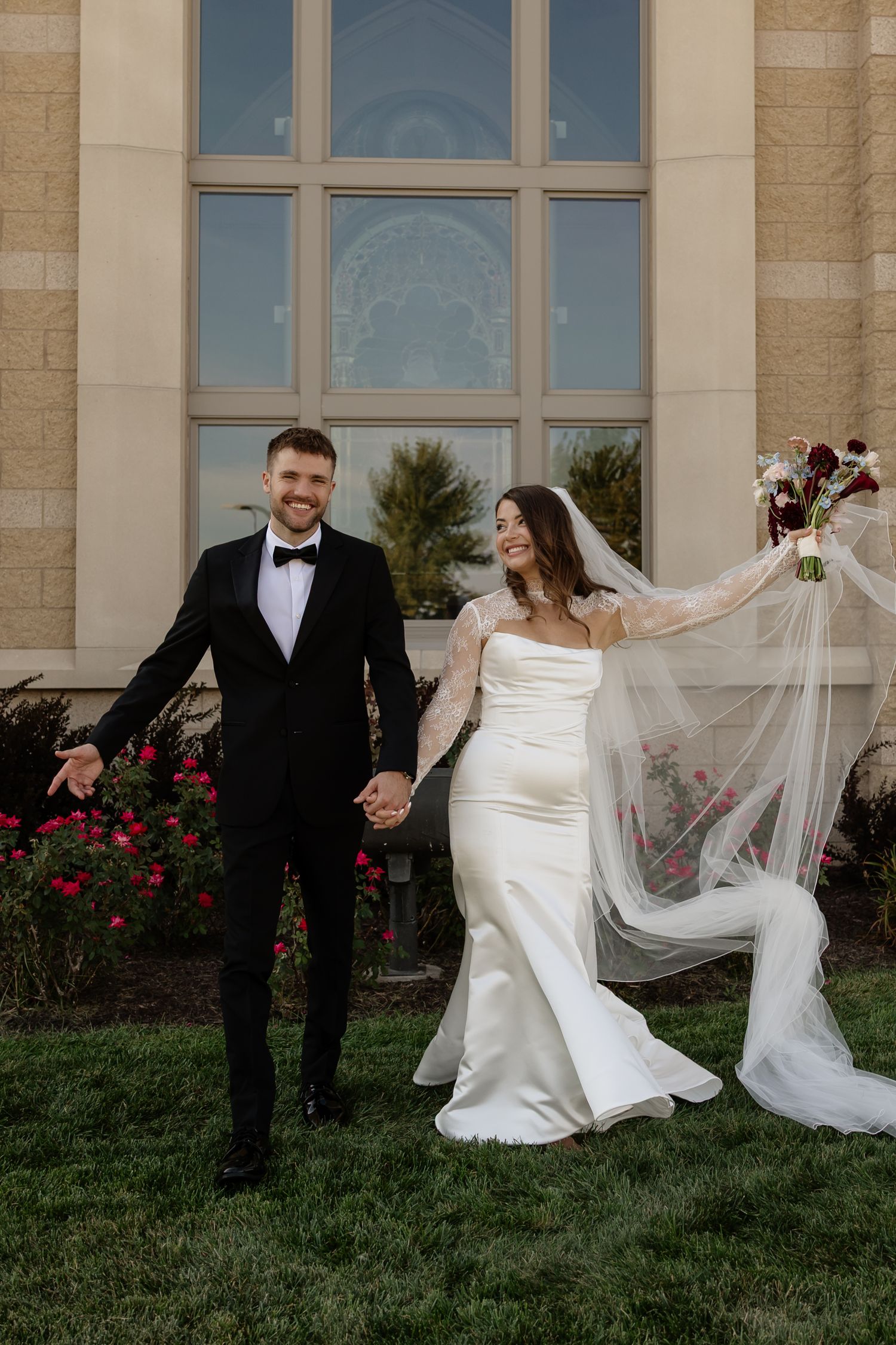 Happy bride and groom holding hands outside a church, bride's veil flowing in the wind, groom in black tuxedo.