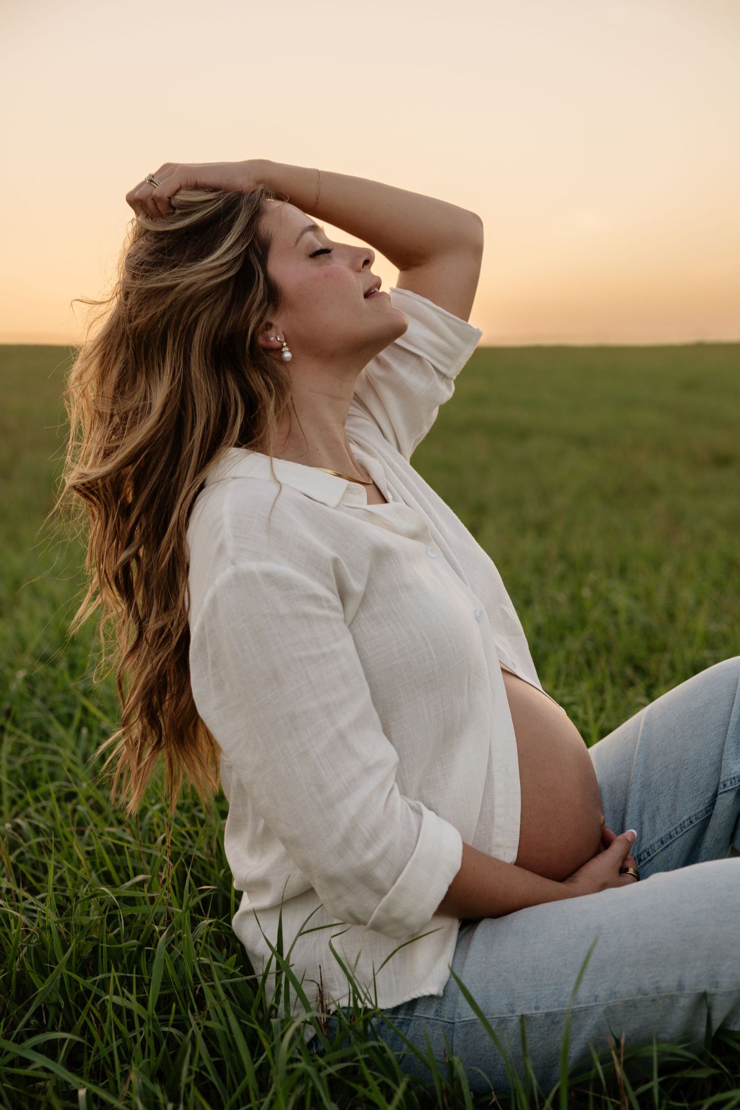 Pregnant woman sitting in green field at sunset, wearing white shirt and jeans, hand in long hair.
