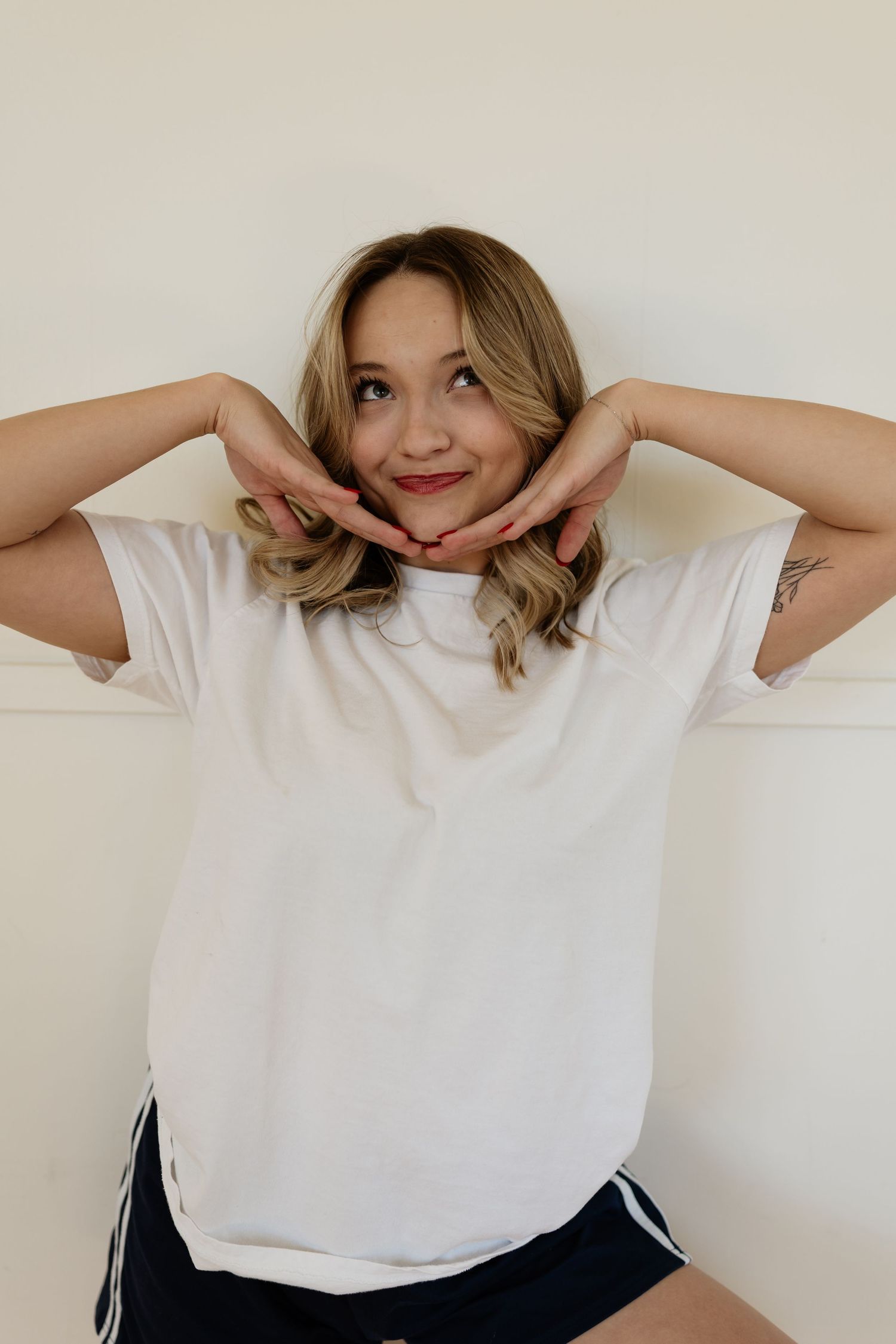 A person in a white t-shirt and dark shorts poses playfully with hands framing their face against a light background.